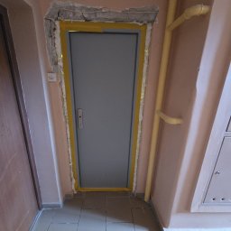 MAX-BUD JAKUB ZARZYŃSKI - Szare drzwi wejściowe, osadzone w ościeżnicy z pianką montażową i żółtą taśmą malarską, widoczne na tle różowej ściany i szarej podłogi z płytek.