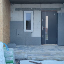 MAX-BUD JAKUB ZARZYŃSKI - Ocieplanie elewacji domu jednorodzinnego z szarym styropianem, widoczne okna i drzwi wejściowe, częściowo zakończone prace, worki z materiałami budowlanymi na pierwszym planie.