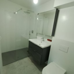 MAX-BUD JAKUB ZARZYŃSKI - Nowoczesna łazienka po remoncie: kabina prysznicowa typu walk-in, szara szafka z umywalką, lustro z szafką, podwieszana toaleta. Minimalistyczny design, jasne płytki.