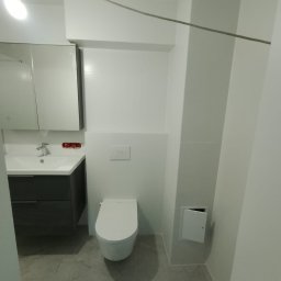 MAX-BUD JAKUB ZARZYŃSKI - Nowoczesna, jasna łazienka po remoncie: podwieszana toaleta, szara szafka z umywalką, białe płytki. Minimalistyczny design z widoczną instalacją elektryczną pod sufitem.