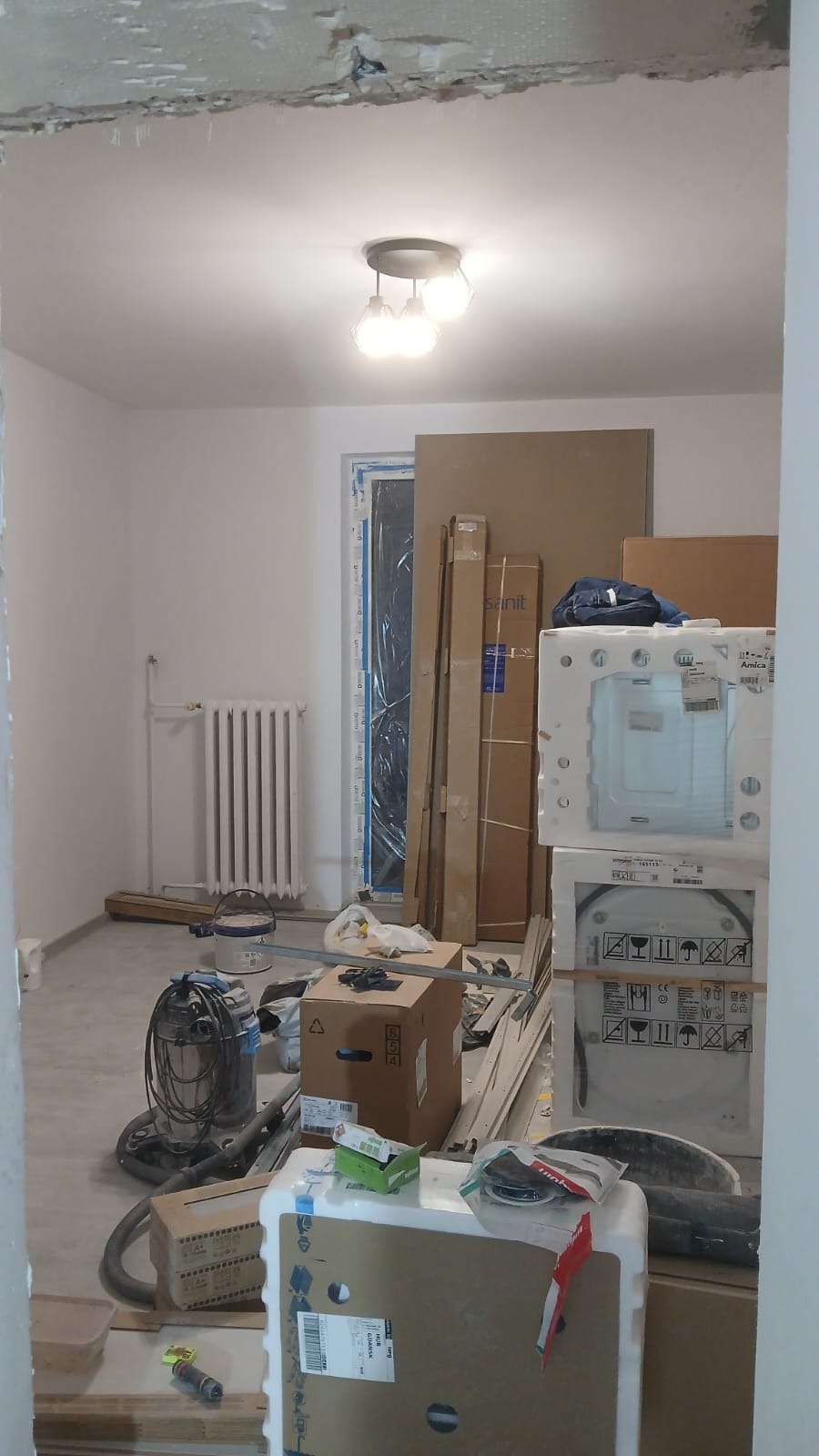 Wnętrze w trakcie prac wykończeniowych: białe ściany, grzejnik, okno w folii, kartony z wyposażeniem łazienki, lampa sufitowa. Widok zza framugi.