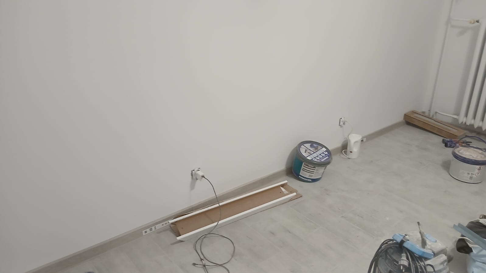 Świeżo pomalowane na biało ściany w rogu pokoju z widocznymi puszkami farby, listwami przypodłogowymi i grzejnikiem. Podłoga wyłożona panelami w odcieniach szarości.