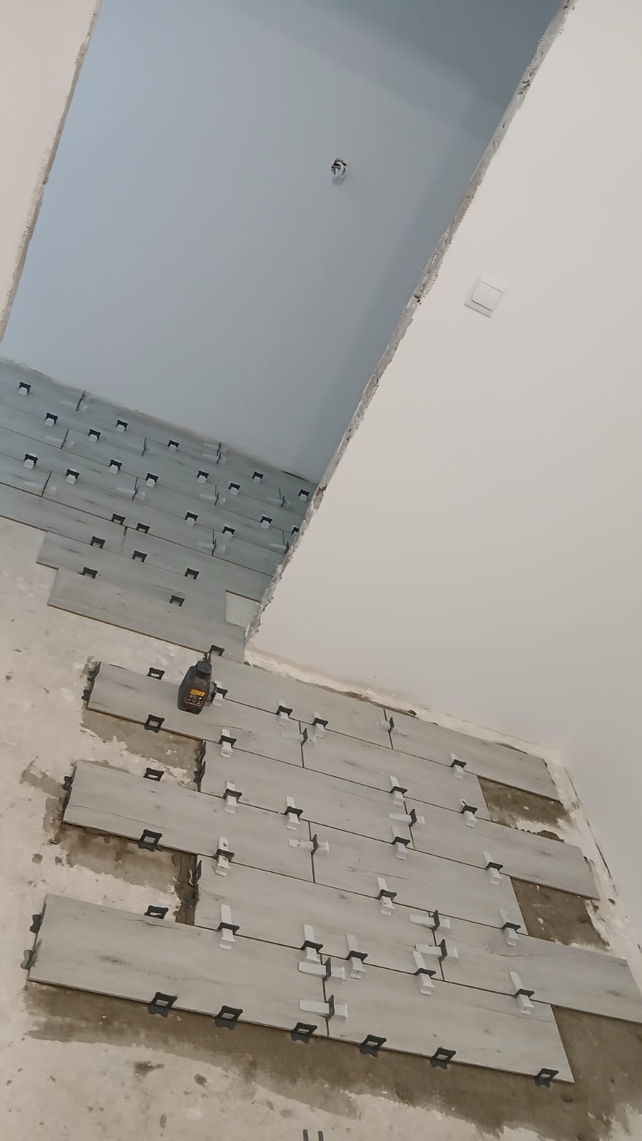 Układanie płytek podłogowych w jasnym odcieniu drewna, z użyciem klipsów poziomujących. Widoczny fragment ściany i sufitu. Ujęcie z góry, pod kątem.