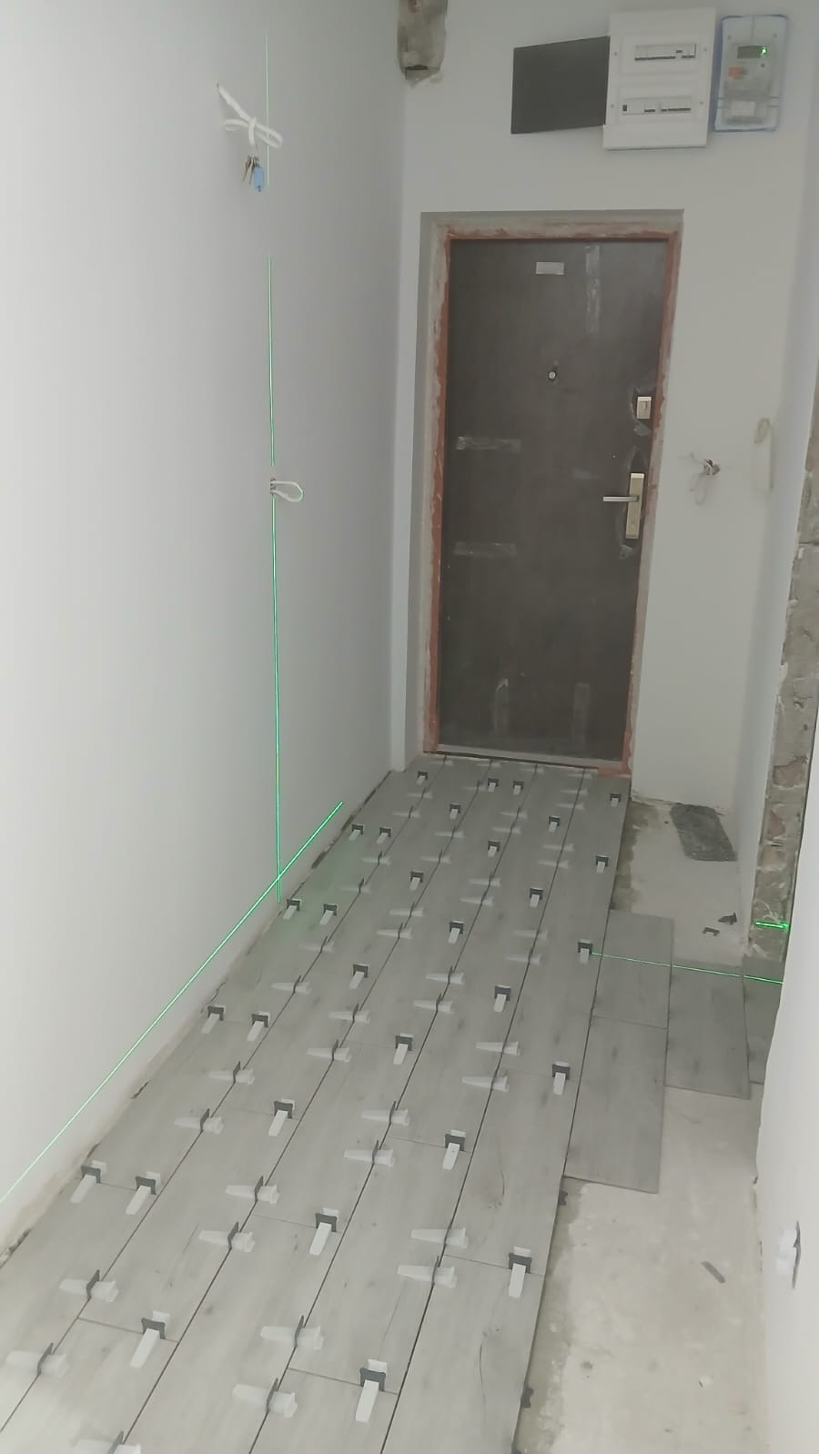 Układanie płytek podłogowych w przedpokoju z użyciem lasera krzyżowego i klipsów poziomujących. Widoczne płytki imitujące drewno i surowy stan ścian.