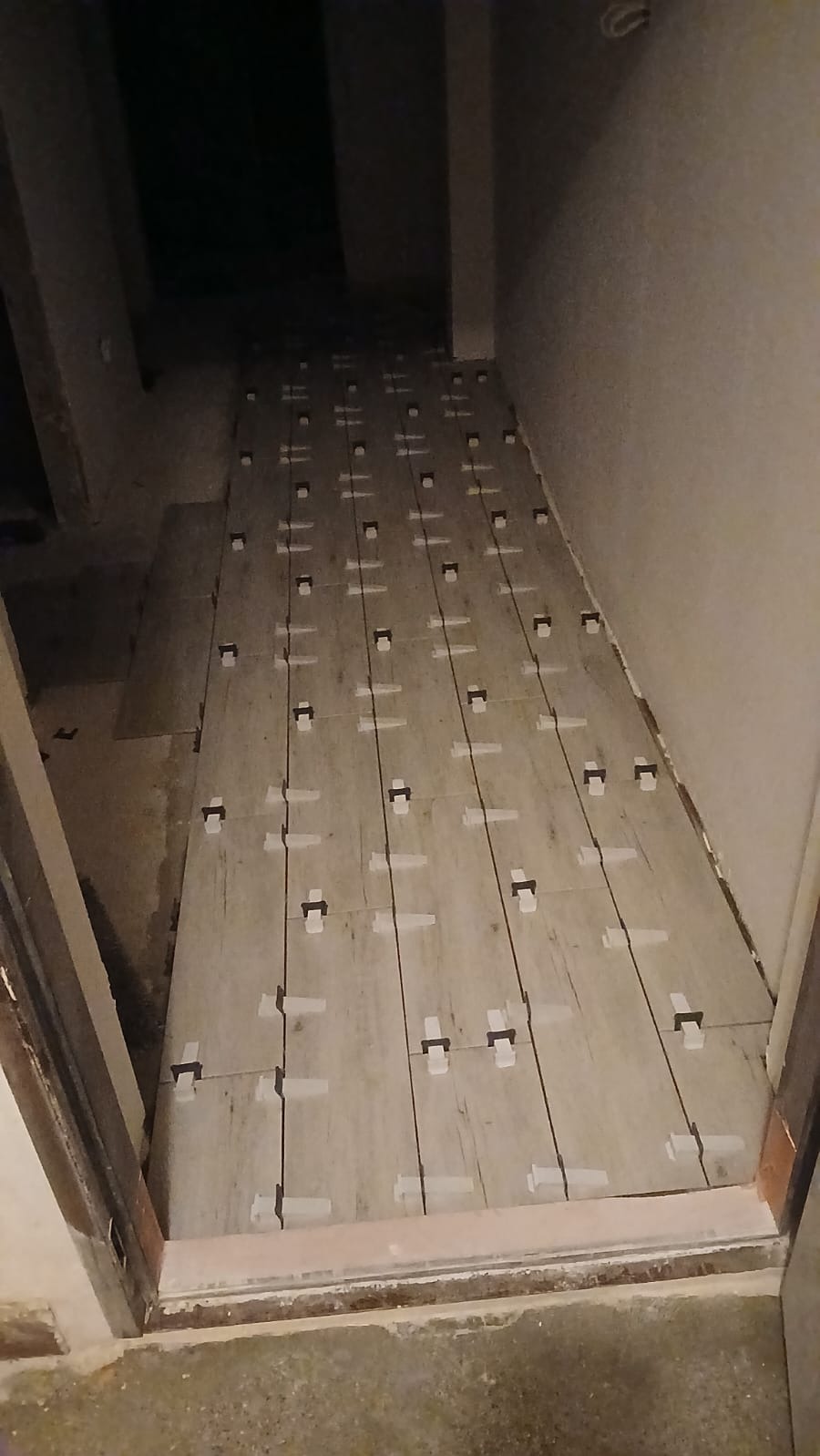 Ułożone płytki podłogowe w korytarzu, z użyciem systemu poziomowania. Widoczne krzyżyki dystansowe i kliny. Ujęcie z perspektywy wejścia do pomieszczenia.