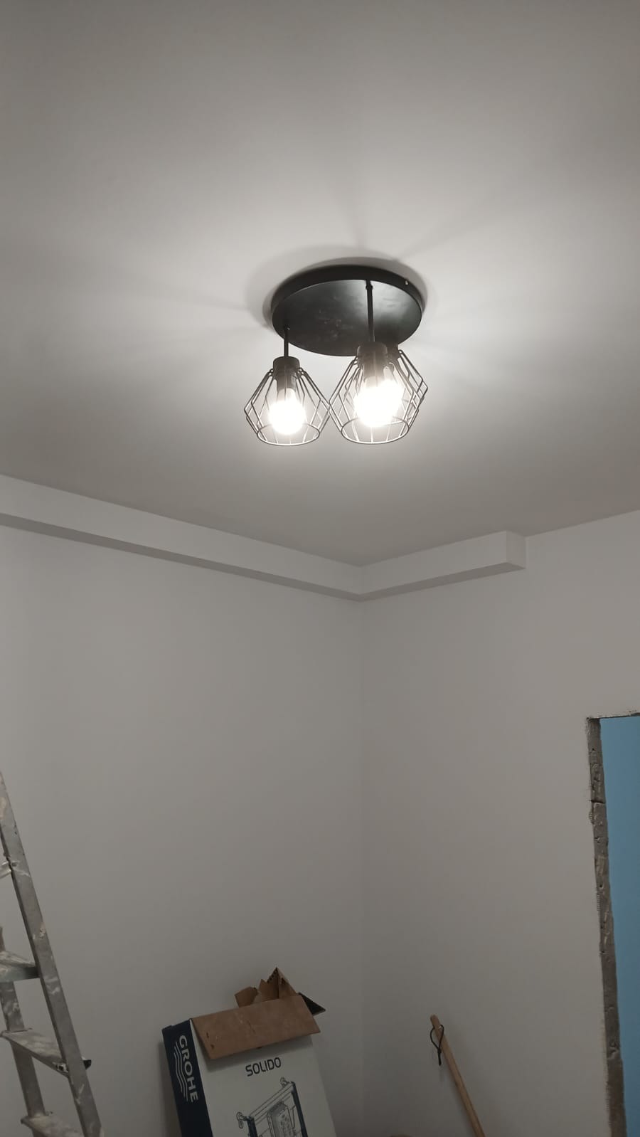 Nowoczesna lampa sufitowa z dwoma żarówkami w metalowych kloszach, zamontowana na białym suficie z dekoracyjną listwą. Widoczny fragment drabiny i karton.