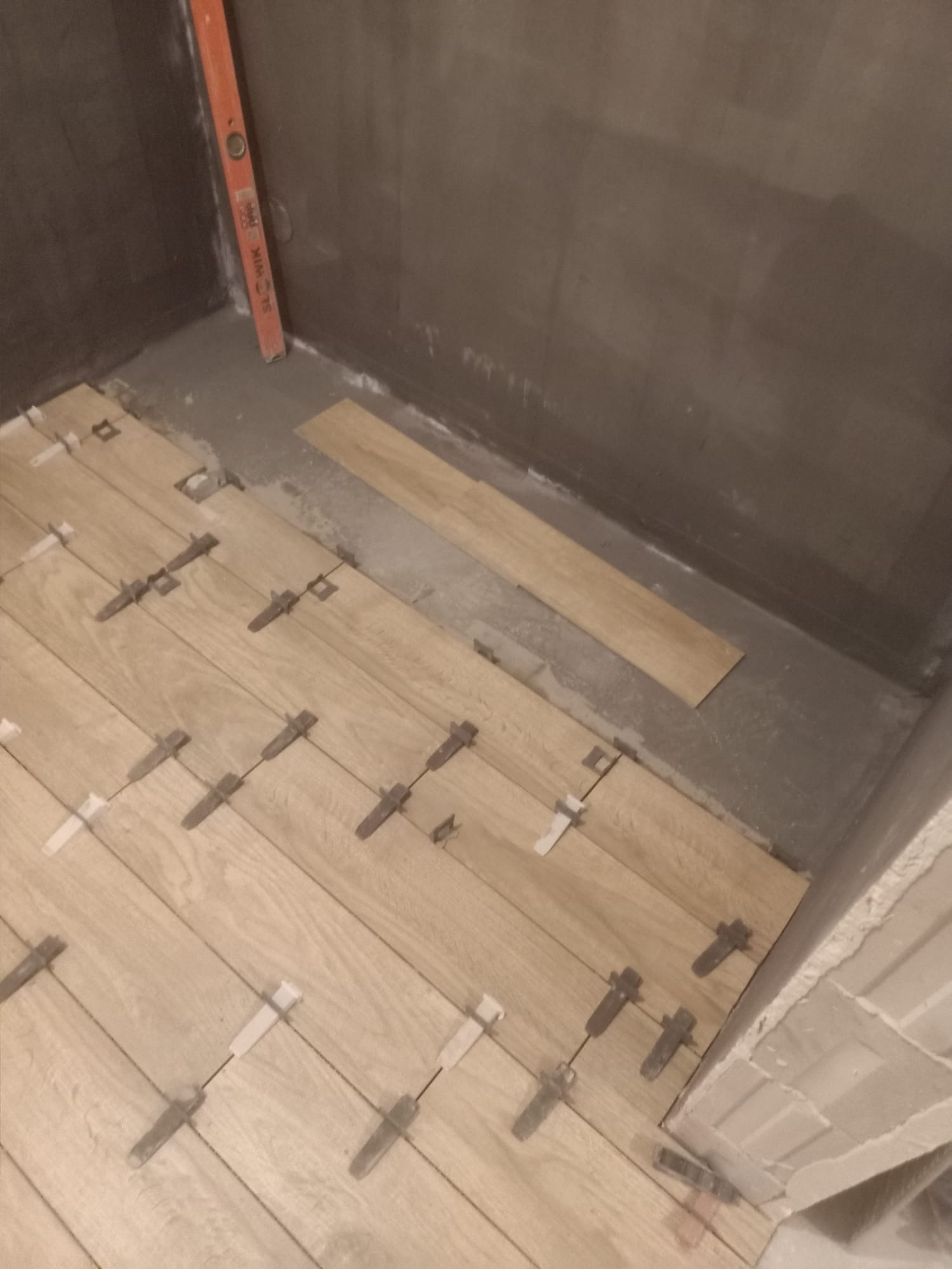 Układanie drewnopodobnych płytek podłogowych w łazience z użyciem klipsów poziomujących. Widoczna poziomica przy ścianie i fragment surowej ściany.