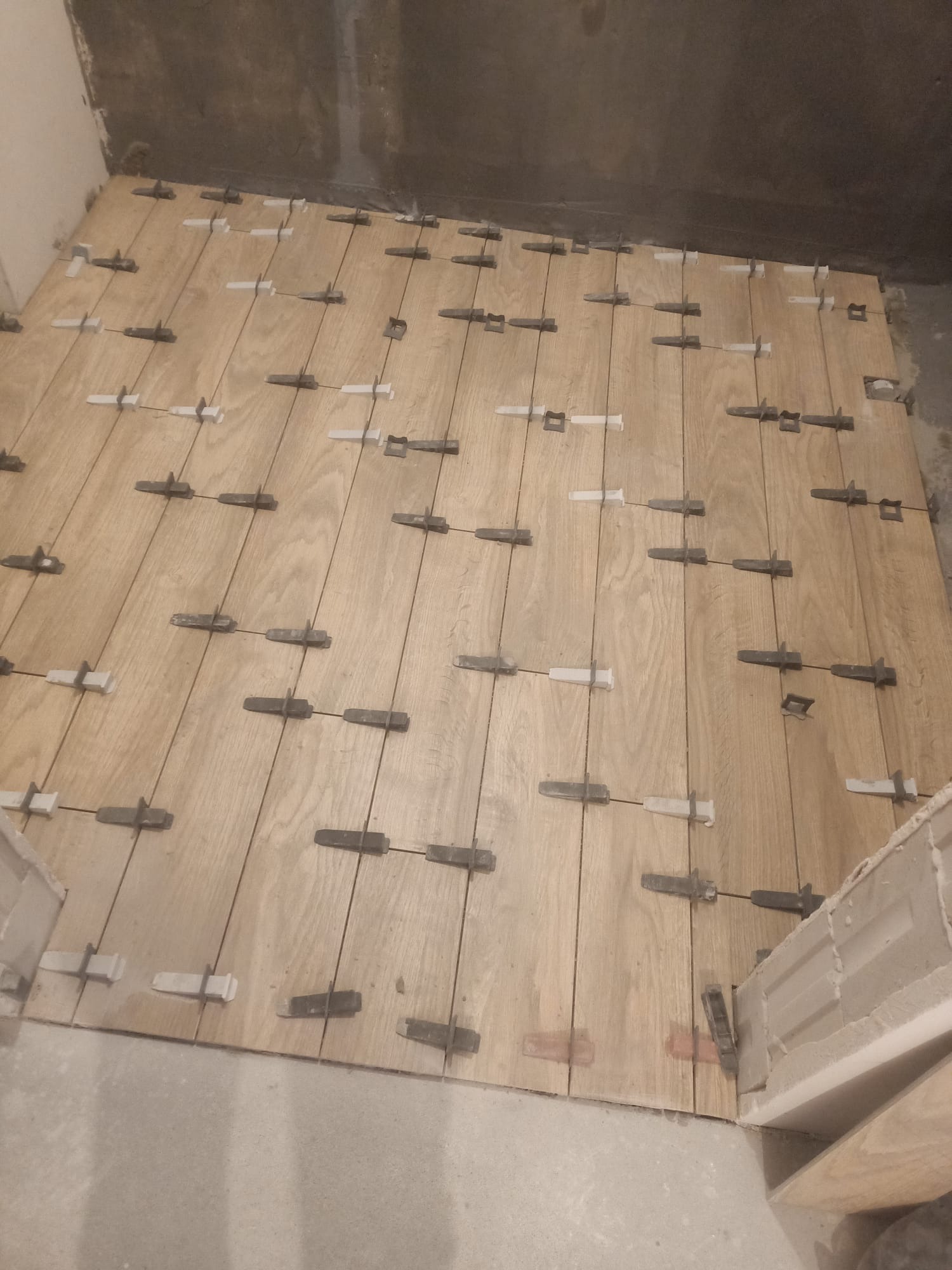 Ułożone płytki podłogowe imitujące drewno, z użyciem klipsów poziomujących, w trakcie prac wykończeniowych. Widoczne surowe ściany i fragment progu.