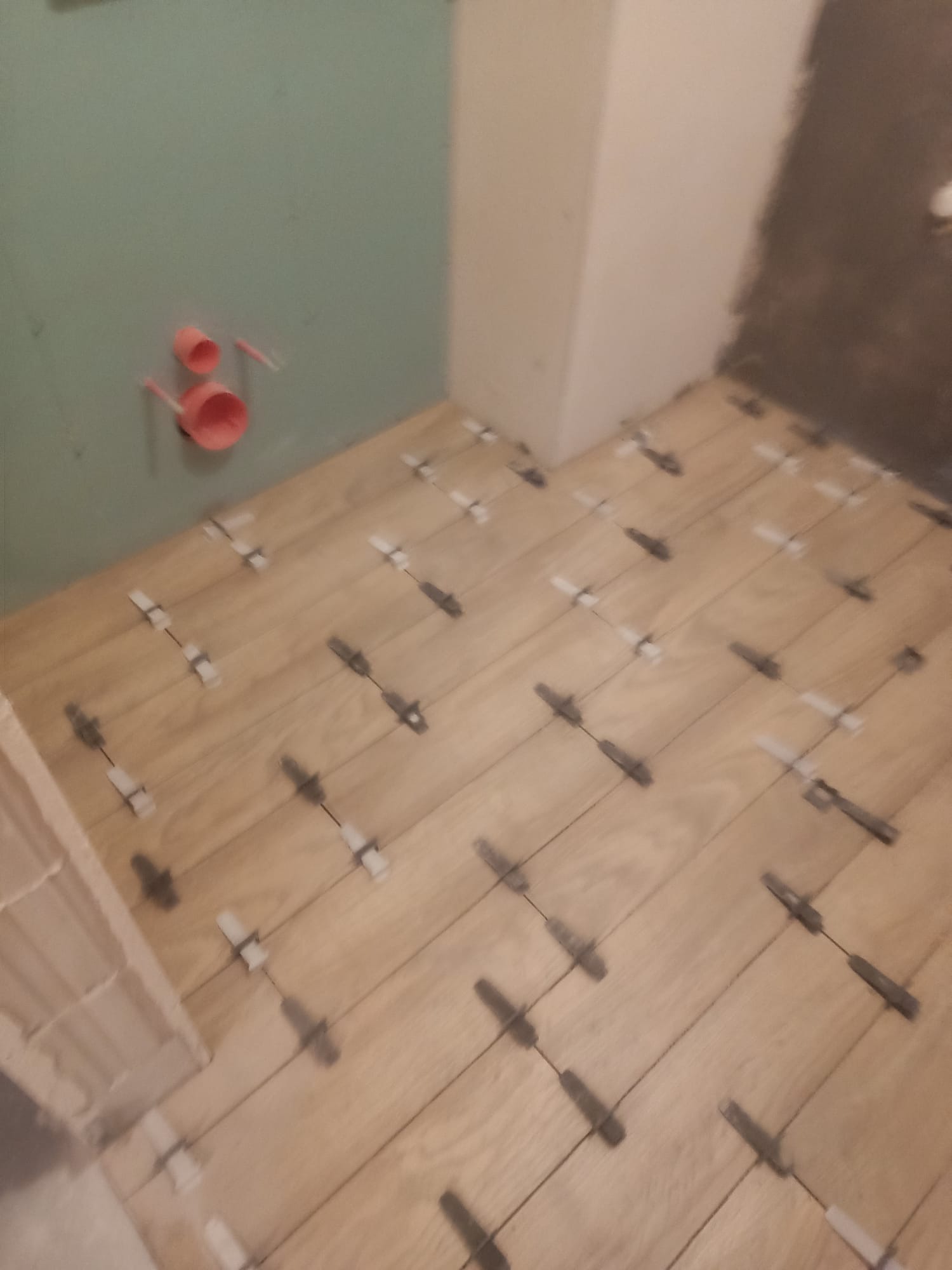 Ułożone płytki podłogowe imitujące drewno w łazience, z użyciem klipsów poziomujących. Widoczne rury odpływowe na ścianie i fragment surowej ściany.