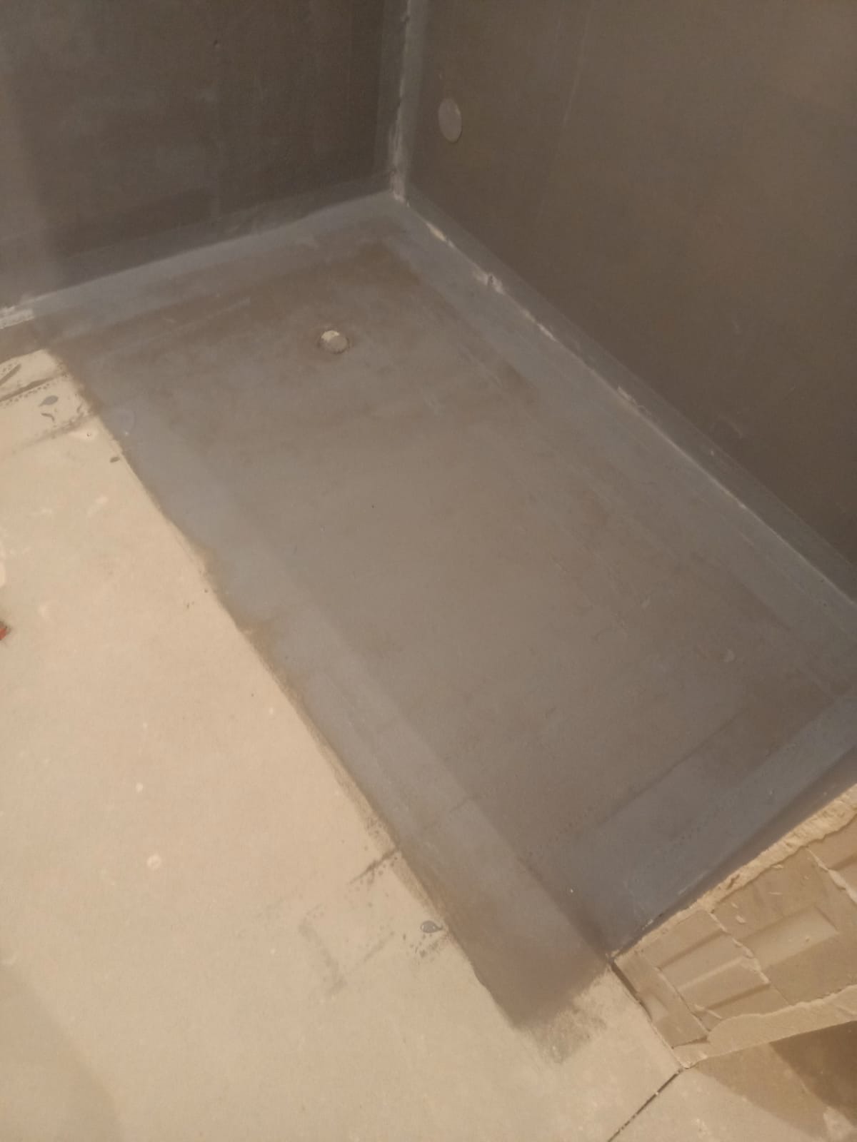 Szara hydroizolacja w rogu łazienki, widoczny odpływ. Przygotowanie do dalszych prac wykończeniowych. Surowe ściany i podłoga przed położeniem płytek.