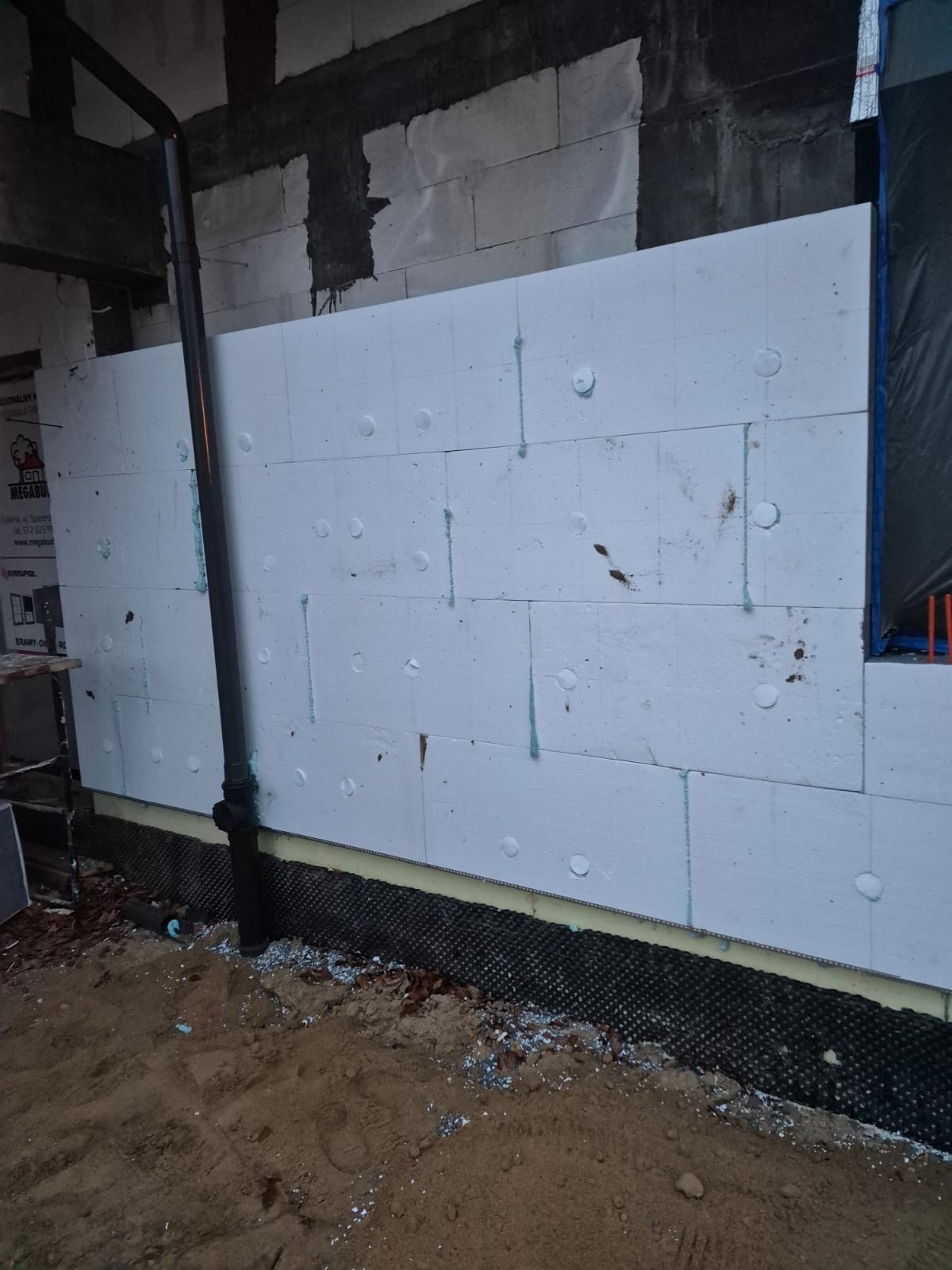 Ocieplanie ściany budynku białym styropianem, widoczna czarna rura spustowa i fragment fundamentu z izolacją. Prace w toku, widoczne ślady kleju i zabrudzenia.