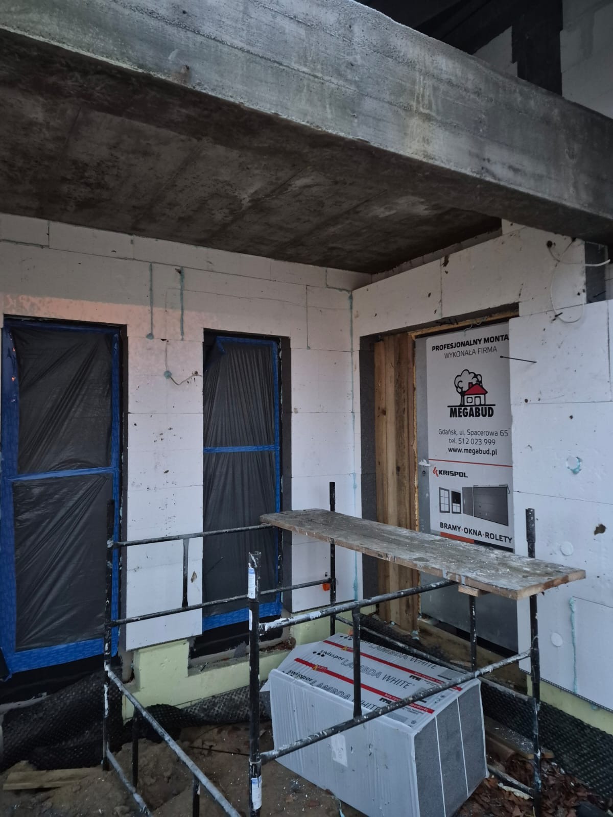 Budowa domu w trakcie: ściany z białych bloczków, okna zabezpieczone folią, rusztowanie i materiały budowlane. Widoczny betonowy strop i reklama firmy Megabud.