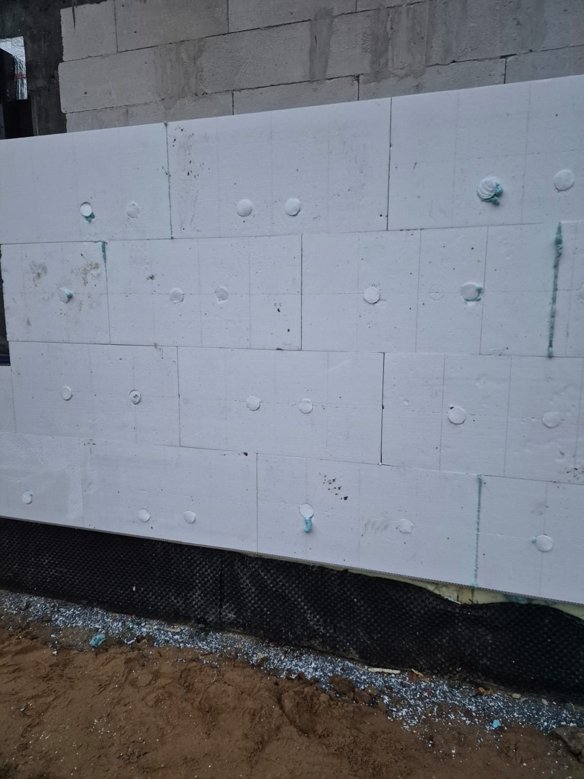 Ocieplanie ściany budynku styropianem, widoczne kołki mocujące i fragment ściany z bloczków. Prace w toku, widoczna czarna folia izolacyjna przy gruncie.