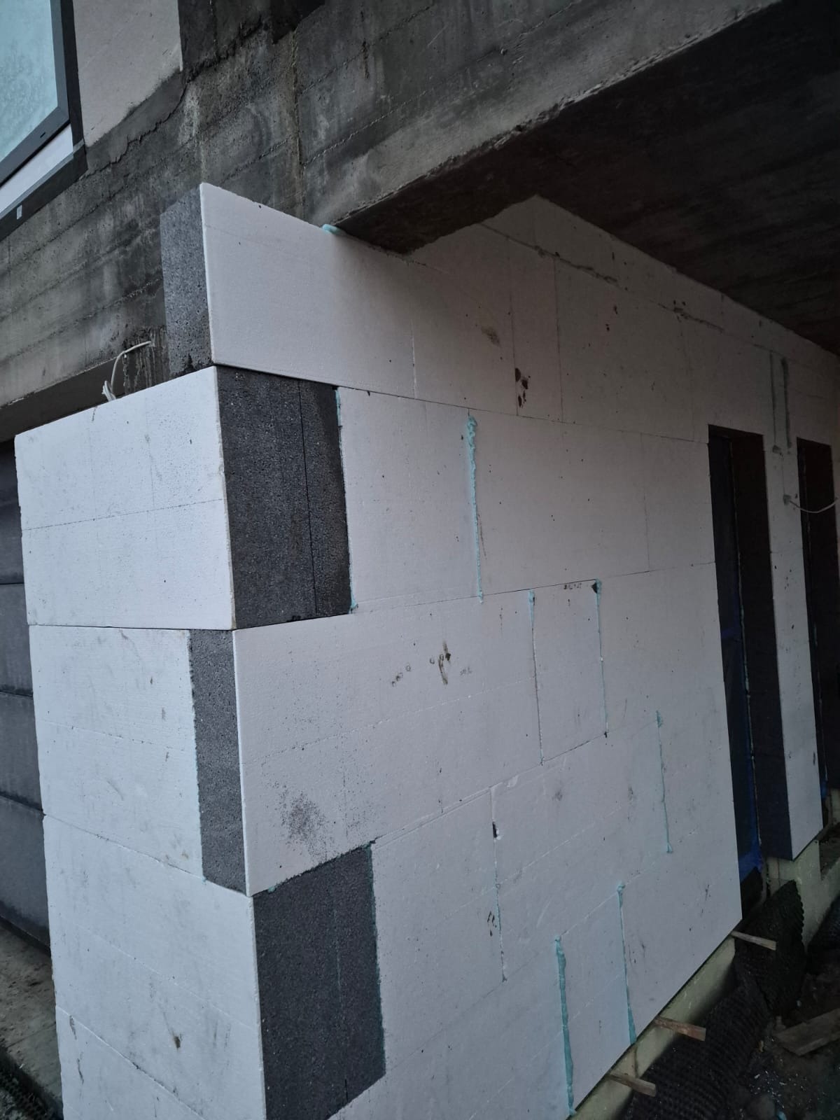 Ściana z białych bloczków z szarymi wstawkami, widoczne spoiny i fragmenty instalacji. Budowa ściany w trakcie realizacji, surowy stan.