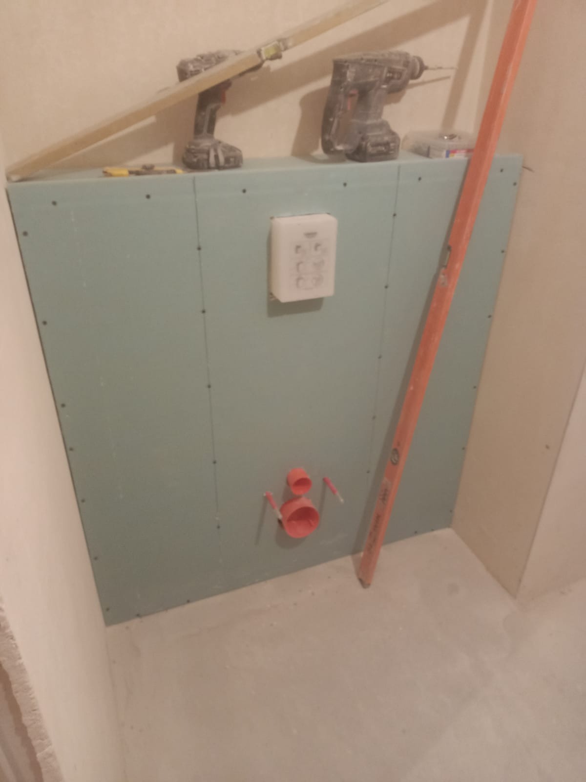 Stelaż podtynkowy zabudowany płytą GK w łazience, widoczne przyłącza wodne i odpływ, poziomica oparta o ścianę, w tle wiertarki na zabudowie. Etap prac wykończeniowych.