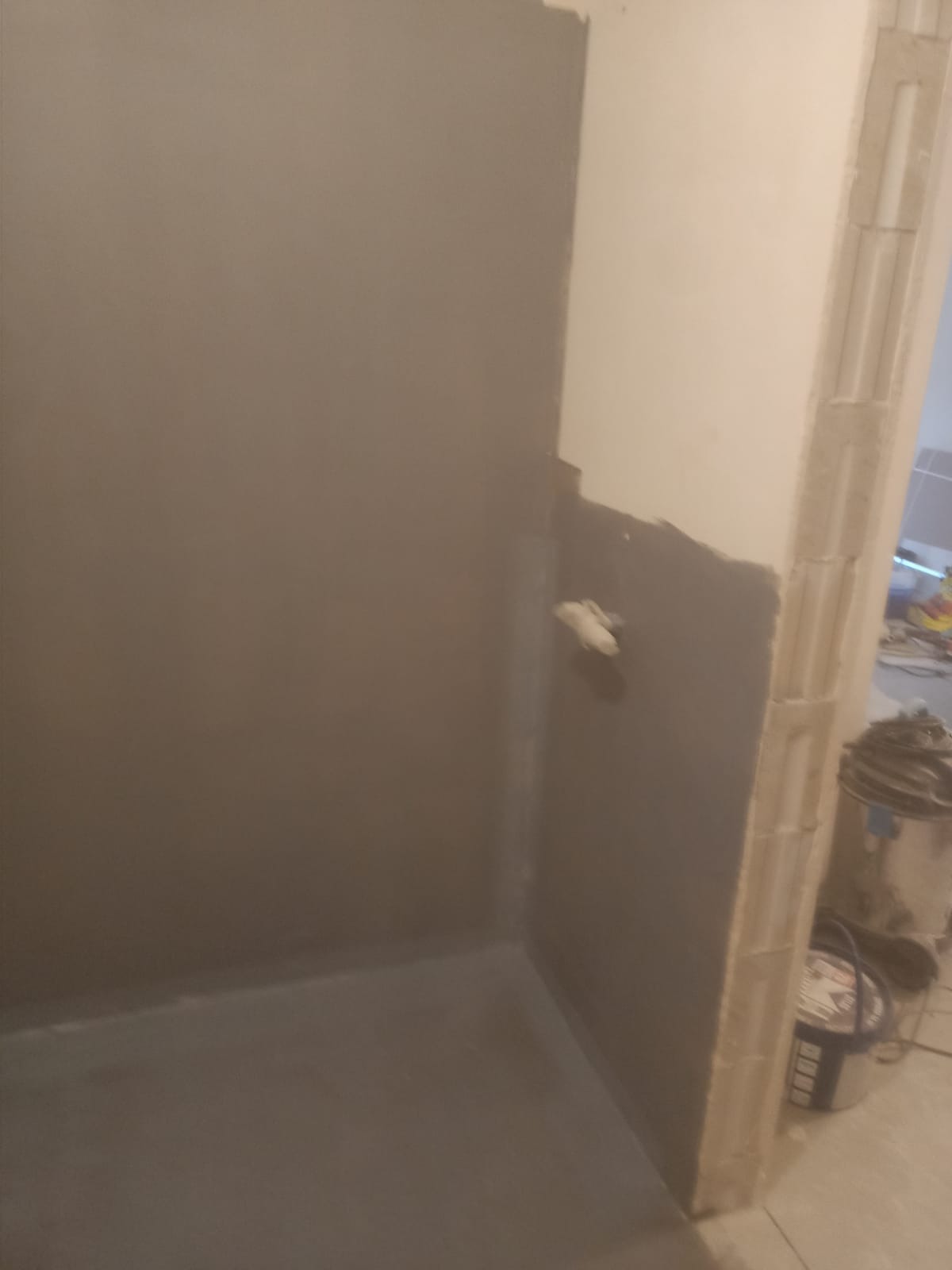 Szara, świeżo nałożona hydroizolacja w rogu łazienki, widoczny fragment ściany z instalacją i wiadro farby w tle. Przygotowanie do dalszych prac wykończeniowych.