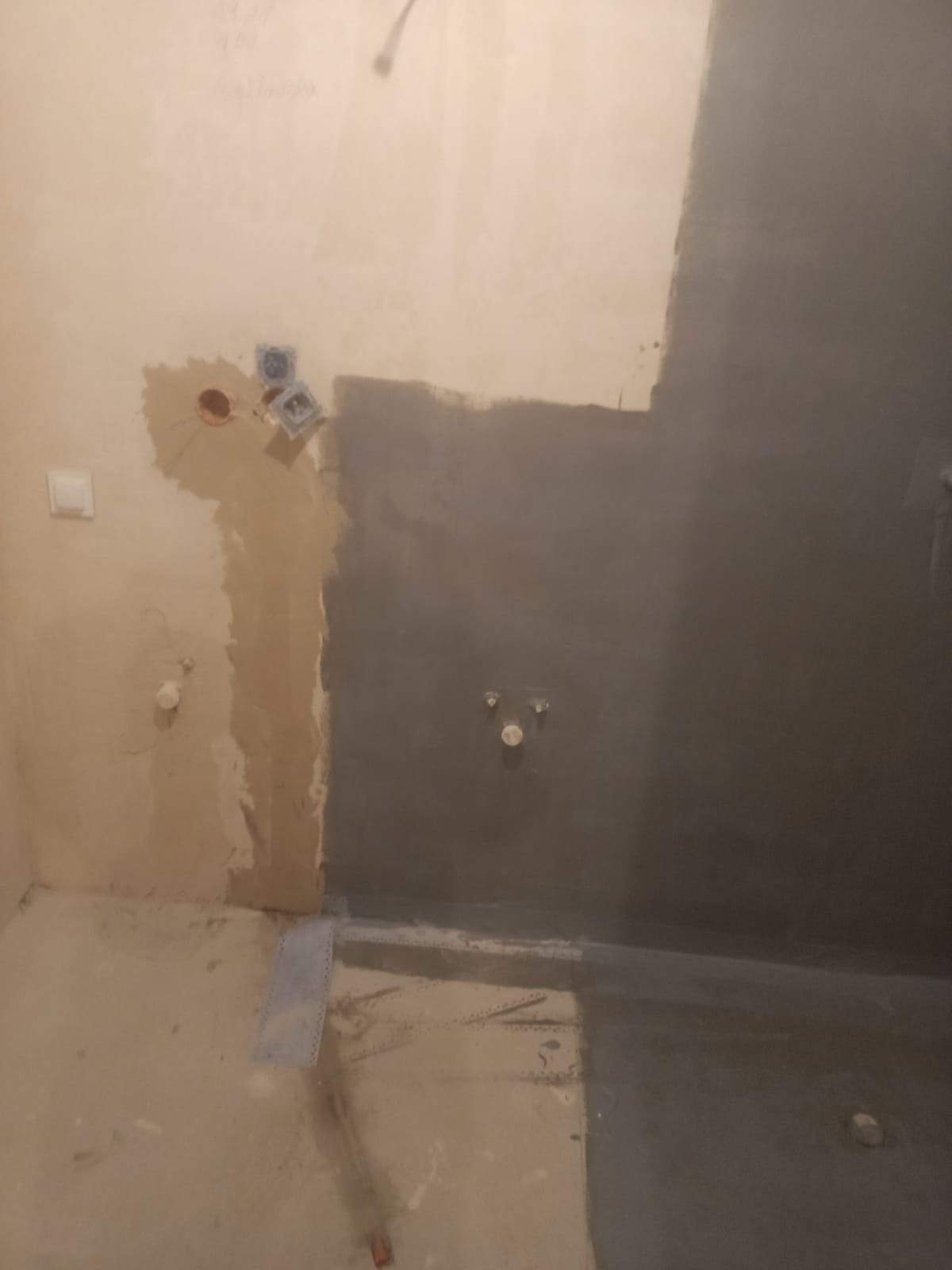 Przygotowanie ścian pod płytki w łazience: fragment ściany z instalacją elektryczną i wodną, częściowo pokryty szarą hydroizolacją. Widoczny odpływ liniowy w podłodze.