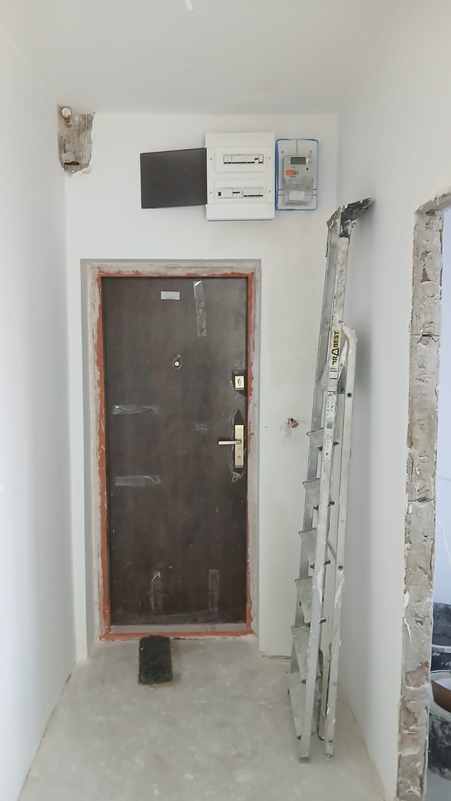 Korytarz w trakcie prac: zabezpieczone drzwi wejściowe, drabina oparta o ścianę, widoczna instalacja elektryczna i fragment uszkodzonego tynku. Etap przygotowania do malowania.