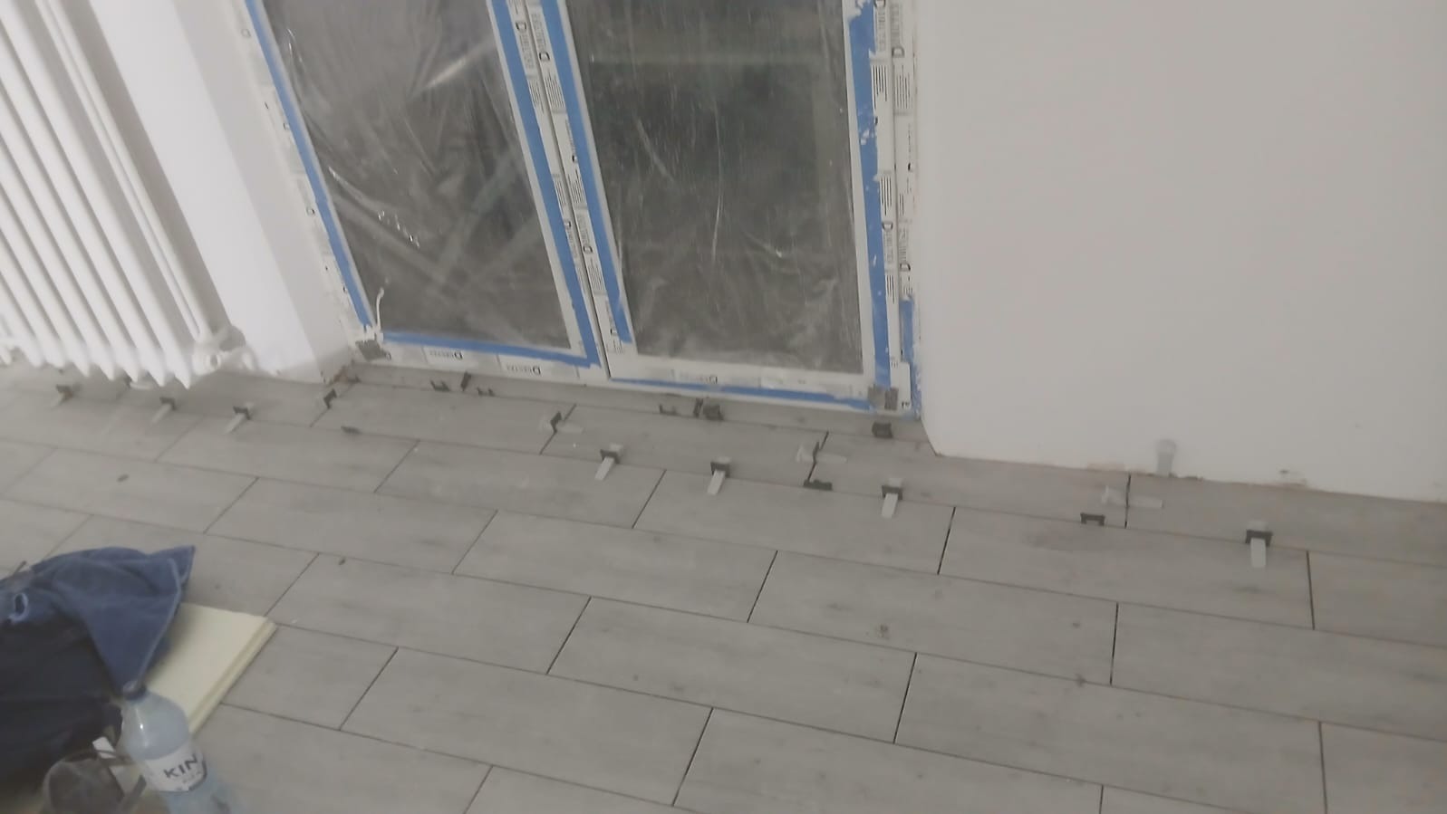 Układanie szarych płytek podłogowych w pokoju, widoczne plastikowe krzyżyki dystansowe, okno zabezpieczone folią. Częściowo widoczny grzejnik i butelka wody.