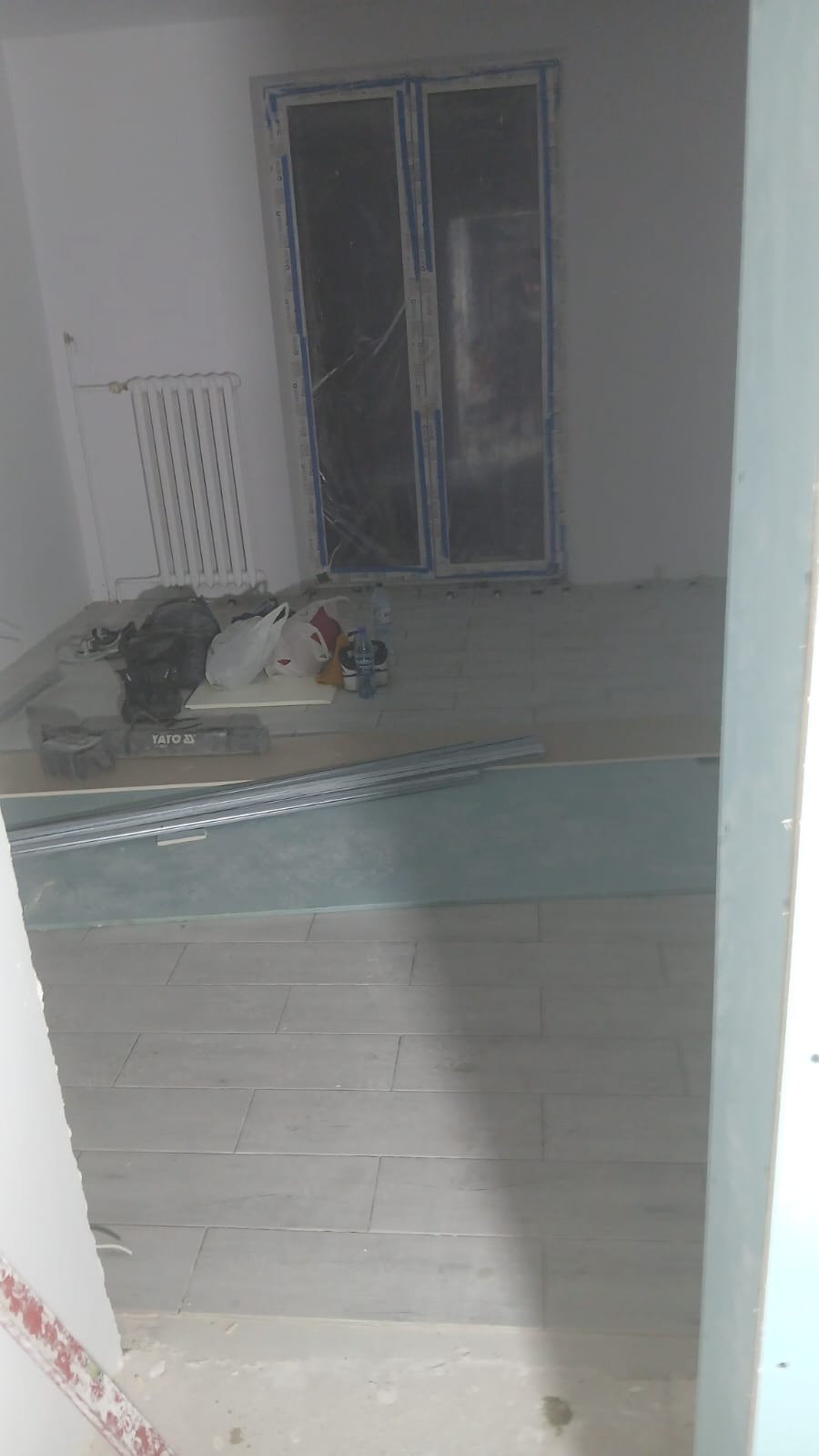 Wnętrze w trakcie prac wykończeniowych: ułożona podłoga z płytek, okno zabezpieczone folią, widoczne narzędzia YATO i profile. Etap prac przed finalnym wykończeniem.