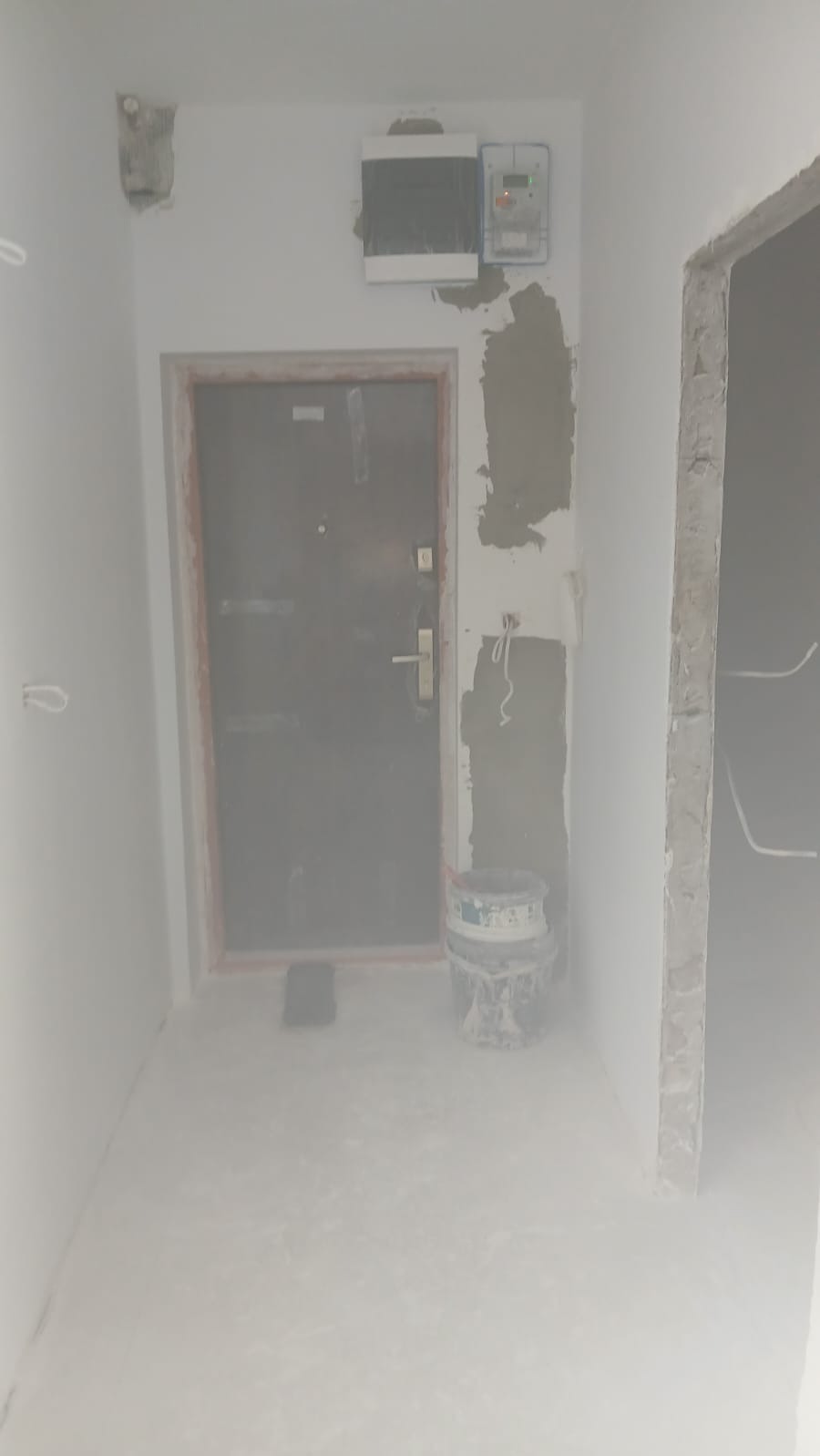 Korytarz w trakcie prac wykończeniowych: szpachlowane ściany, widoczna puszka elektryczna, drzwi wejściowe i wiadro po farbie. Wnętrze w stanie surowym.