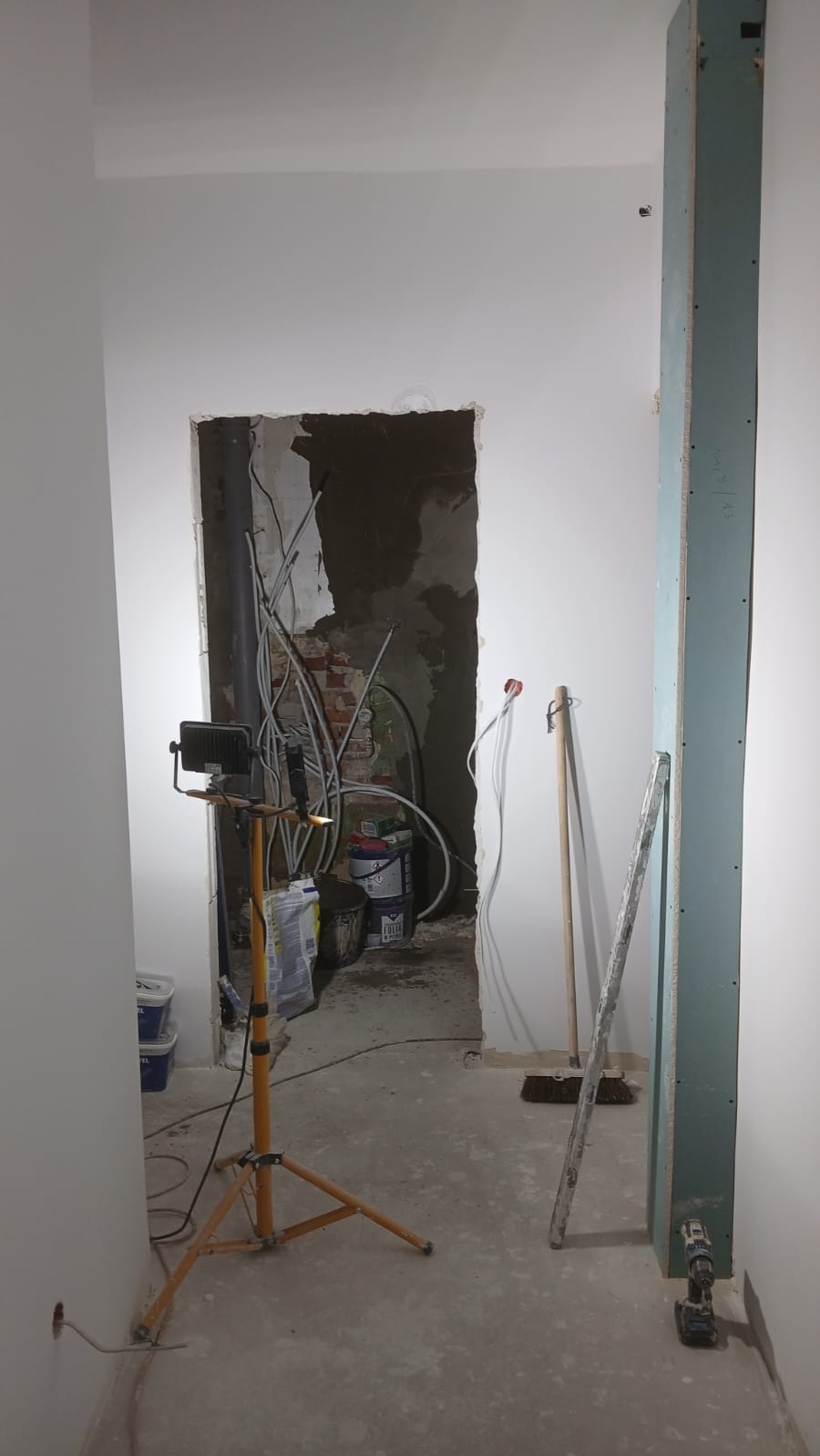Wnętrze w trakcie remontu: odsłonięte przewody, surowe ściany, lampa na statywie i narzędzia. Widoczne przygotowania do dalszych prac wykończeniowych.
