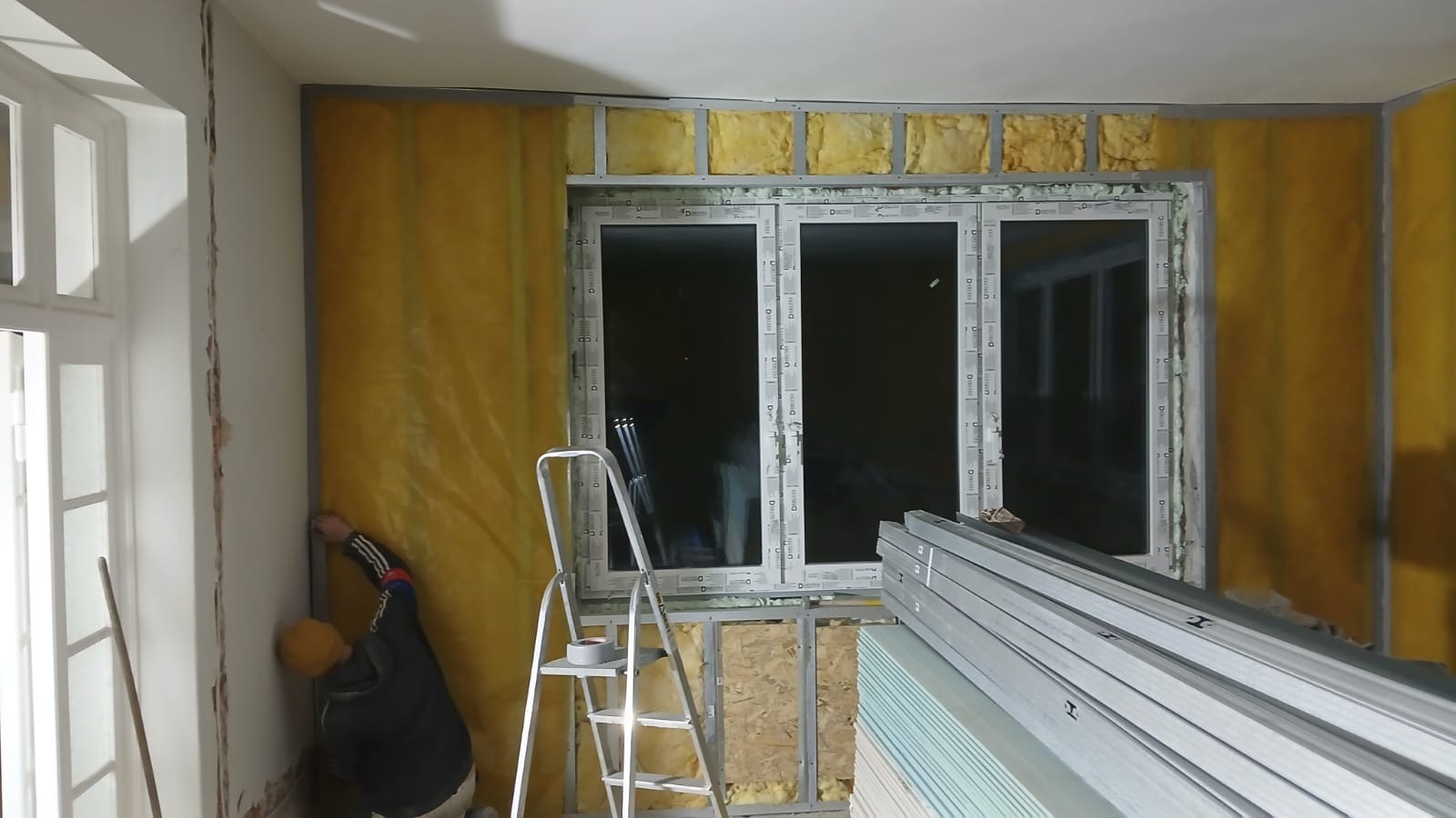 Ocieplanie ścian wełną mineralną wewnątrz pomieszczenia, widoczne profile metalowe, okno PCV i drabina. Prace wykończeniowe w trakcie realizacji.