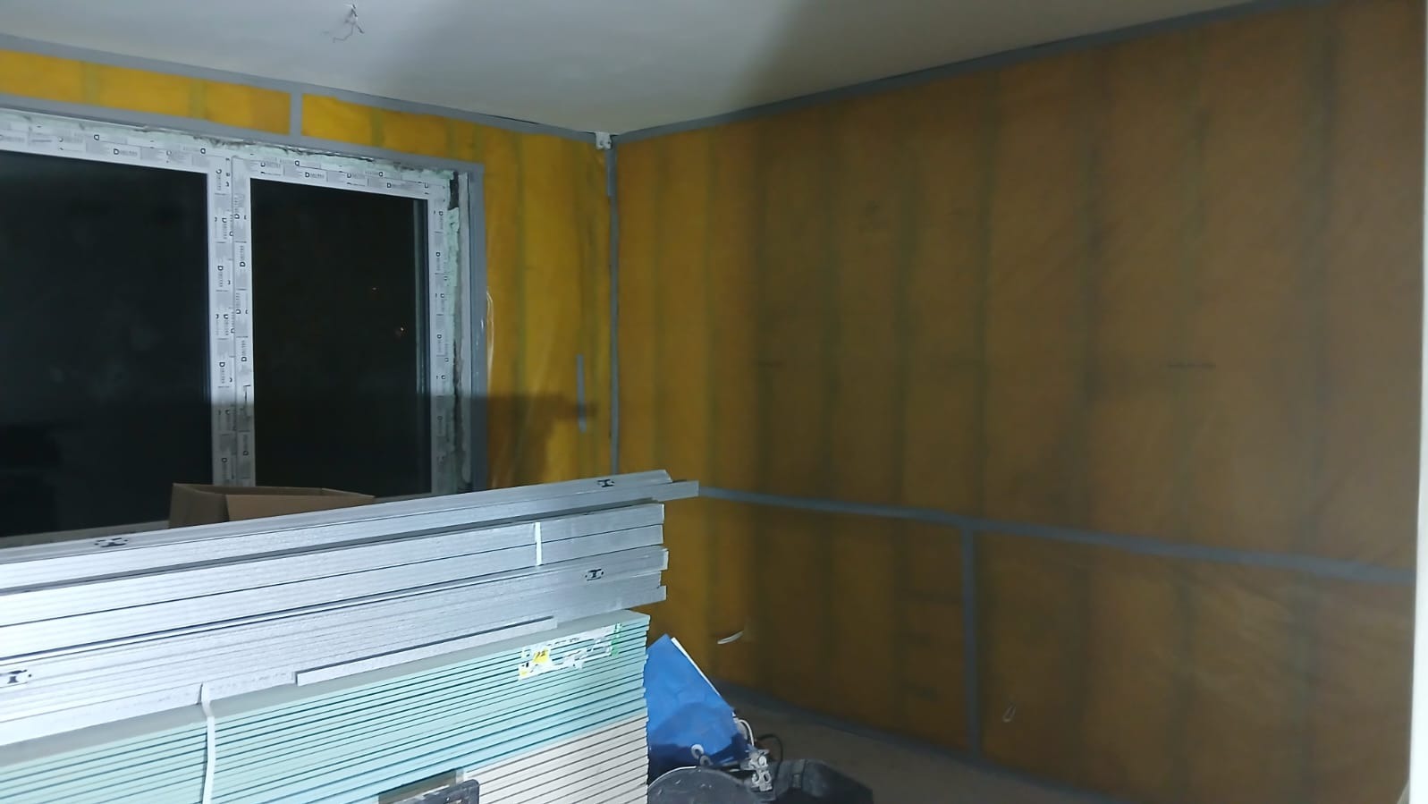 Wnętrze w trakcie ocieplania: żółta wełna mineralna na ścianach, profile i płyty gipsowe przy oknie. Etap prac wykończeniowych, widoczne materiały budowlane.