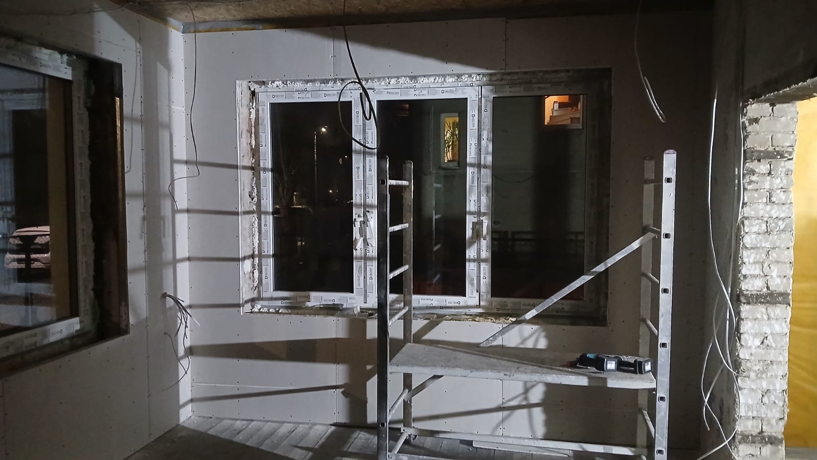 Wnętrze w trakcie prac wykończeniowych: ściany z płyt gipsowo-kartonowych, okna PCV, drabina i narzędzia. Widoczne przewody elektryczne i surowy mur.