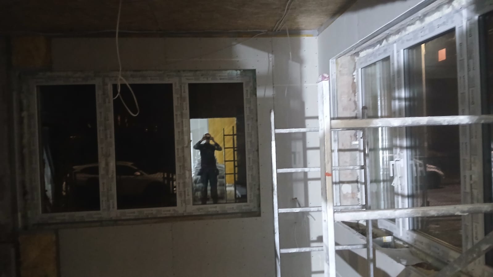 Montaż okien PCV w surowym wnętrzu. Widoczne profile okienne, drabina i odbicie osoby w oknie. Prace wykończeniowe w trakcie realizacji.