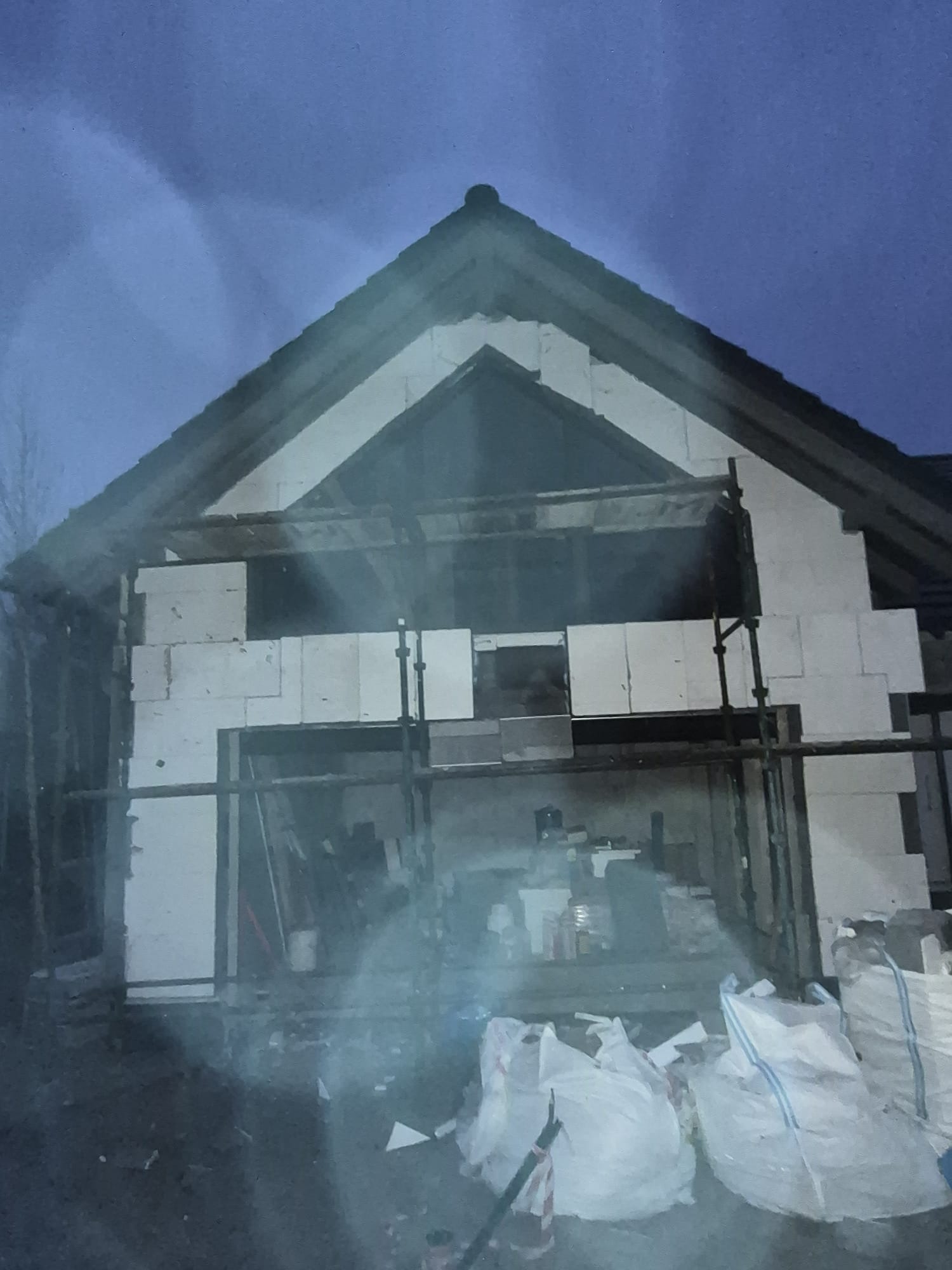 Budowa domu w trakcie: ściany z białych bloczków, rusztowania, worki z materiałami budowlanymi. Widok na szczyt budynku w pochmurny dzień.