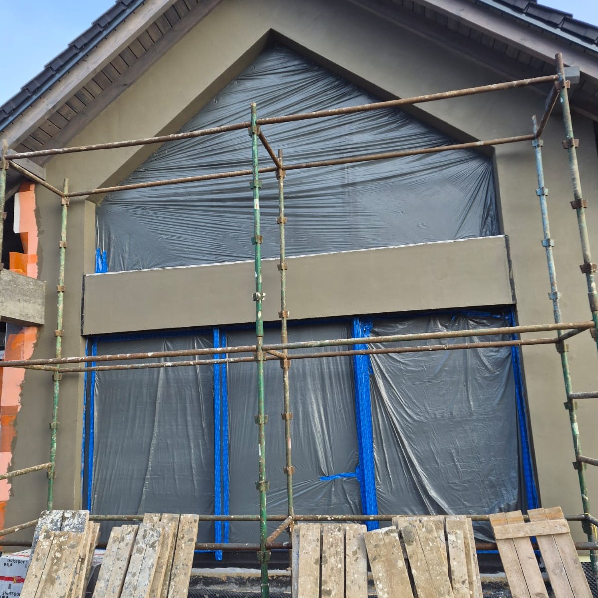 Elewacja domu w trakcie prac: nałożony tynk, rusztowanie, okna zabezpieczone folią. Widoczny fragment ocieplenia. Ujęcie z dołu, w dzień.