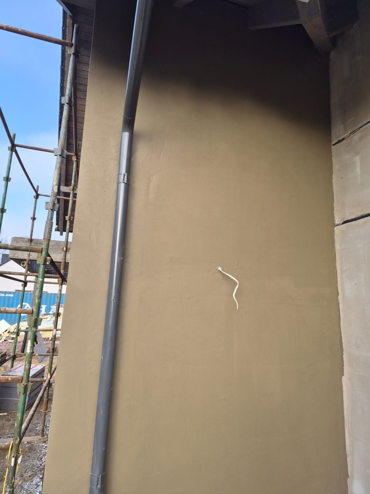 Świeżo otynkowana elewacja budynku w odcieniach beżu, z widoczną rynną spustową i fragmentem rusztowania. Kabel wystaje ze ściany. Ujęcie z dołu, w dziennym świetle.