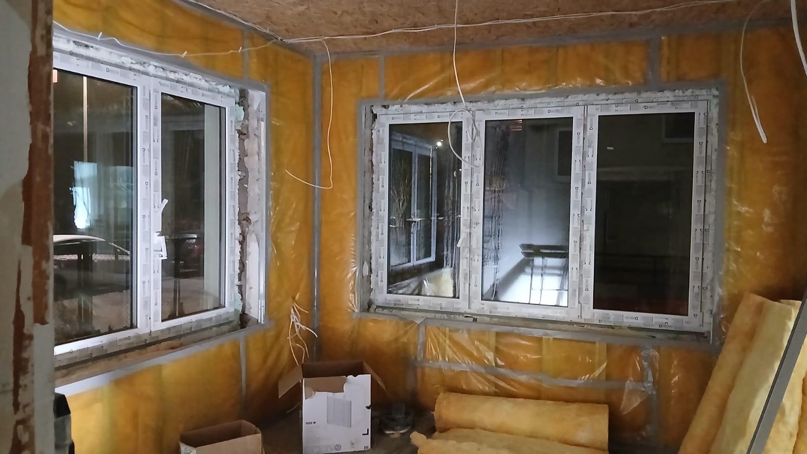 Wnętrze w trakcie ocieplania: żółta folia izolacyjna na ścianach, białe ramy okienne, kartony i rolki wełny mineralnej. Widoczne przewody elektryczne.