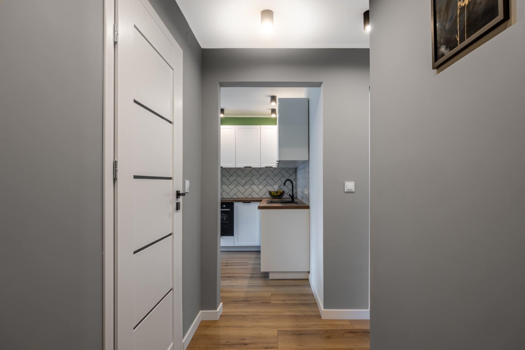 Nowoczesne wnętrze: szara ściana, drewniana podłoga, białe drzwi z czarnymi akcentami i widok na kuchnię z białymi szafkami i zielonym akcentem.