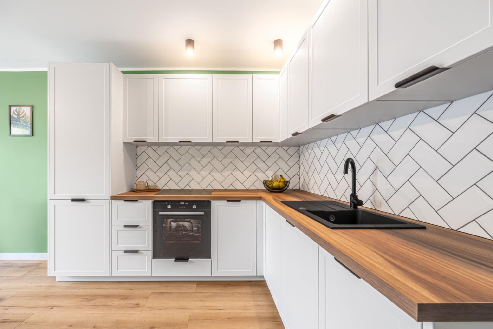 Nowoczesna kuchnia z białymi szafkami, drewnianym blatem i czarną armaturą. Ściana z białych płytek ułożonych w jodełkę. Jasne drewniane podłogi i zielona ściana w tle.