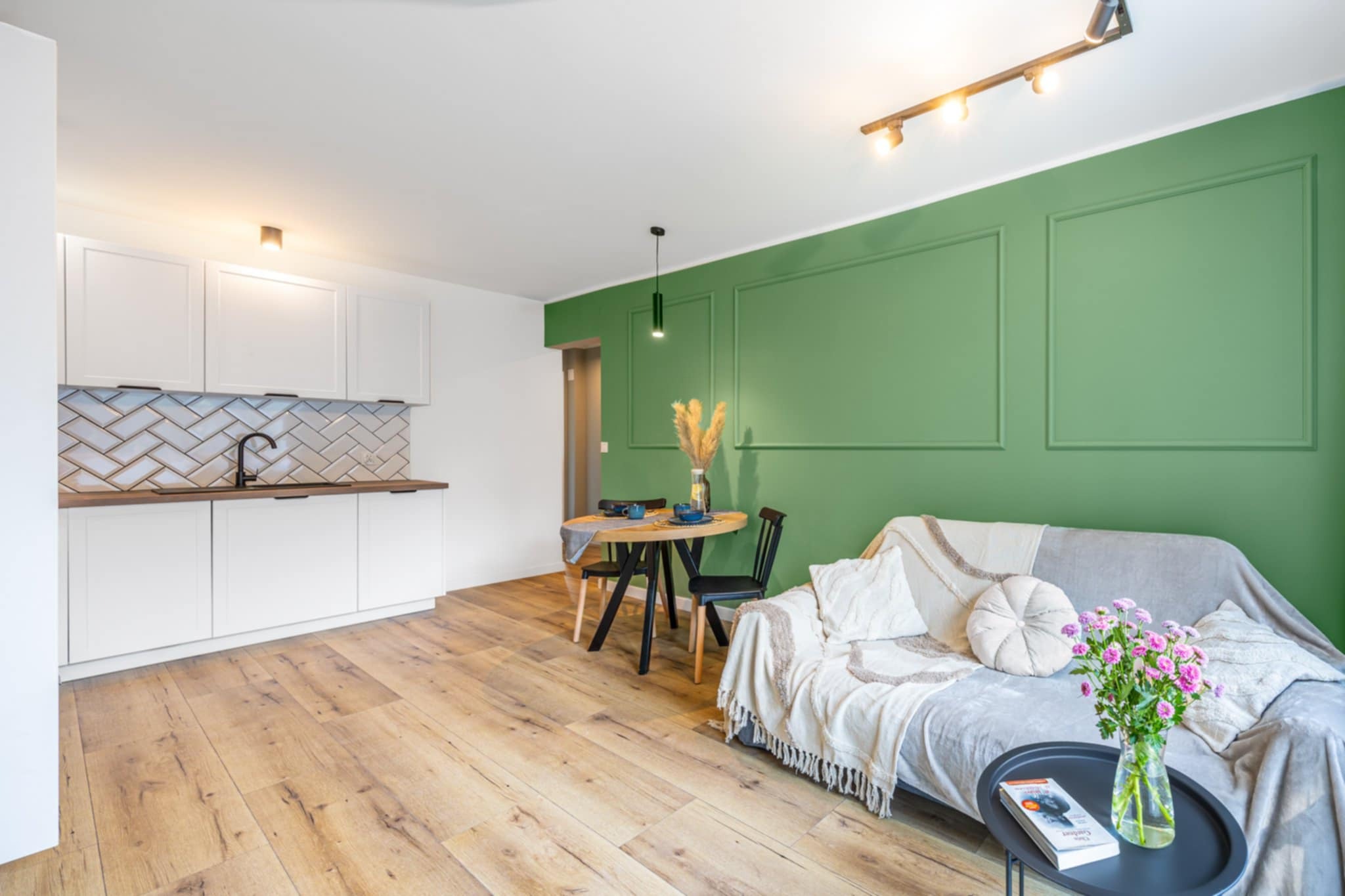 Jasne wnętrze mieszkania po remoncie: drewniana podłoga, biała zabudowa kuchenna z czarną armaturą, zielona ściana z dekoracyjnymi panelami i wygodna sofa.