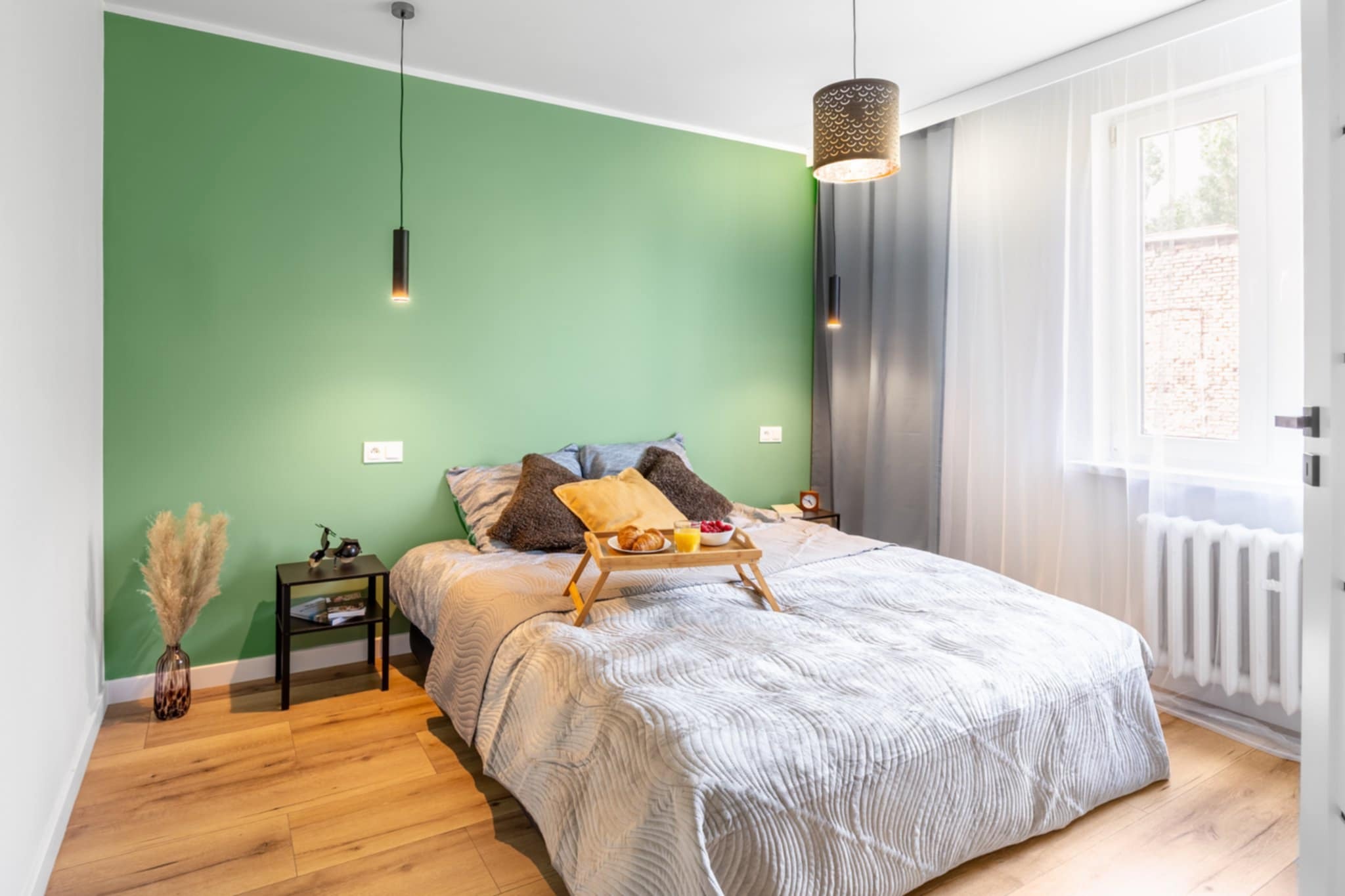 Jasna sypialnia z zieloną ścianą, drewnianą podłogą i łóżkiem z śniadaniem na tacy. Minimalistyczne lampy wiszące i subtelne dekoracje tworzą przytulną atmosferę.