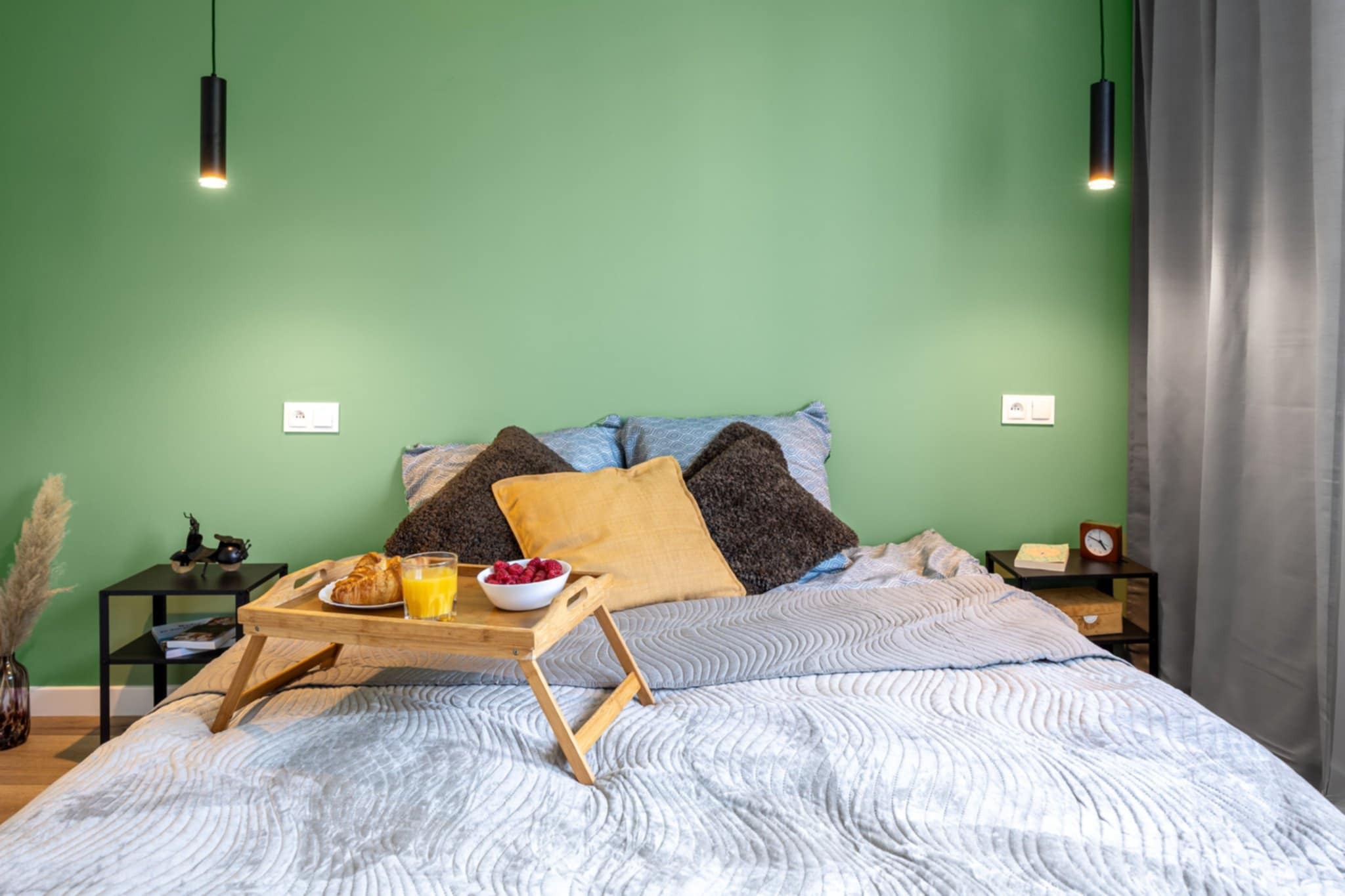 Sypialnia z zieloną ścianą, łóżkiem z szarą narzutą i śniadaniem na tacy. Dwie czarne lampy wiszące po bokach łóżka. Nowoczesny design wnętrza.