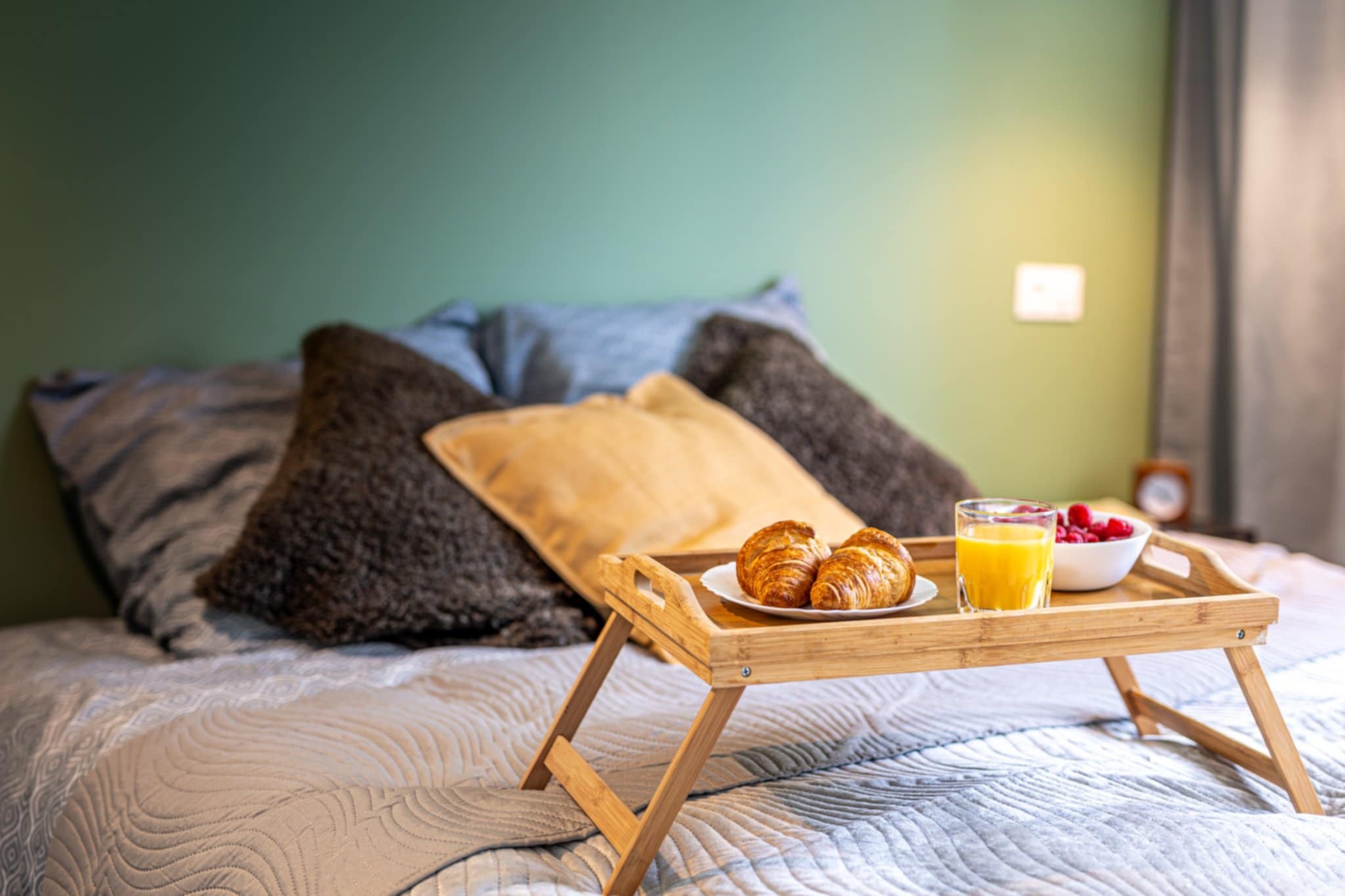 Śniadanie na łóżku: rogaliki, sok pomarańczowy i maliny na drewnianej tacy z nóżkami. W tle poduszki i zielona ściana. Przytulne wnętrze sypialni.