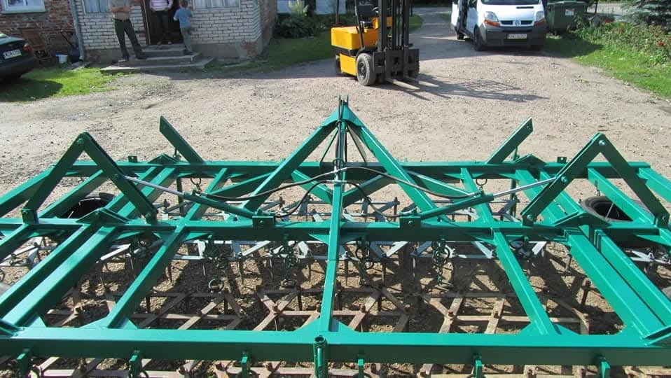 Zielony agregat uprawowy z hydraulicznymi siłownikami, widoczny na podwórku w tle budynek, wózek widłowy i samochód dostawczy.