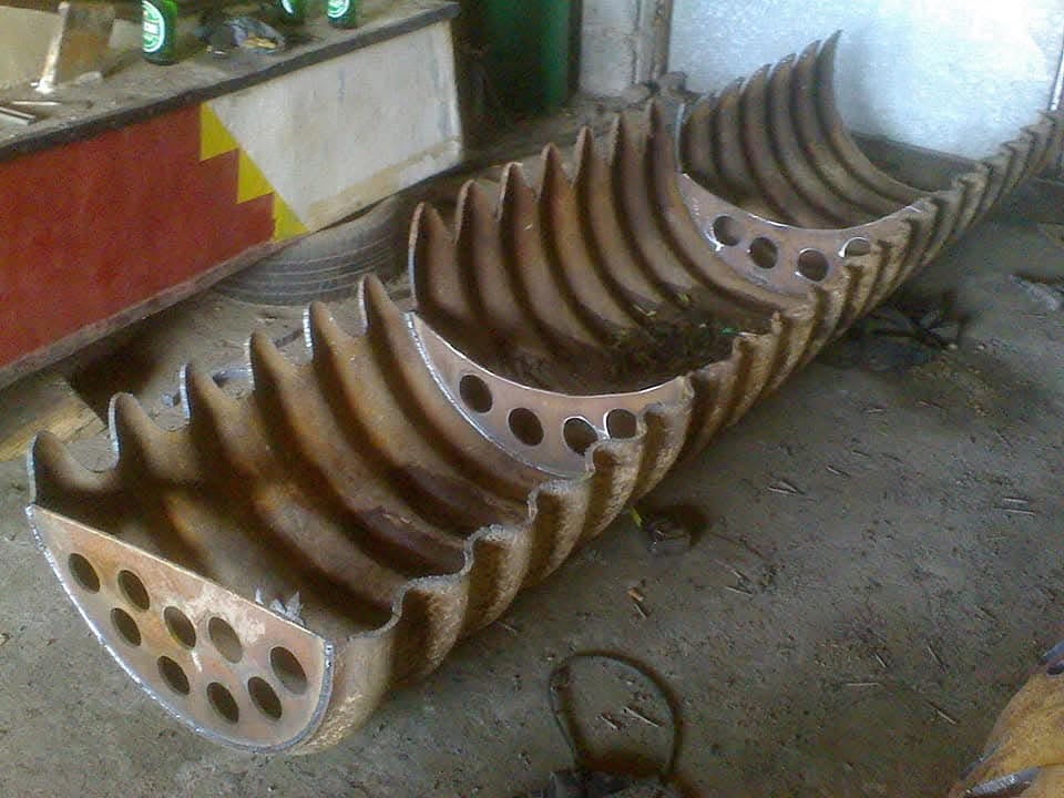 Fragment spawanej konstrukcji metalowej z otworami, leżącej na betonowej podłodze w warsztacie, widoczne ślady spawania i rdzy.