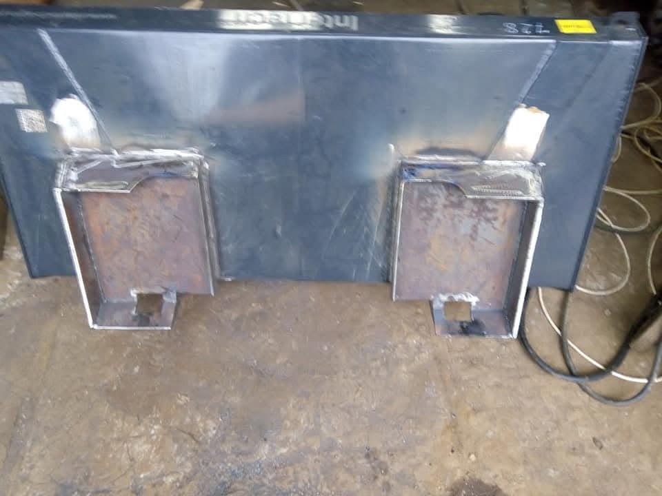 Metalowa konstrukcja z przyspawanymi elementami mocującymi, widoczne spawy i surowa powierzchnia metalu, kabel zasilający w tle.