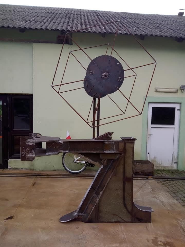 Metalowa rzeźba plenerowa z elementami konstrukcyjnymi, w tle budynek z szarym dachem i flaga Polski.