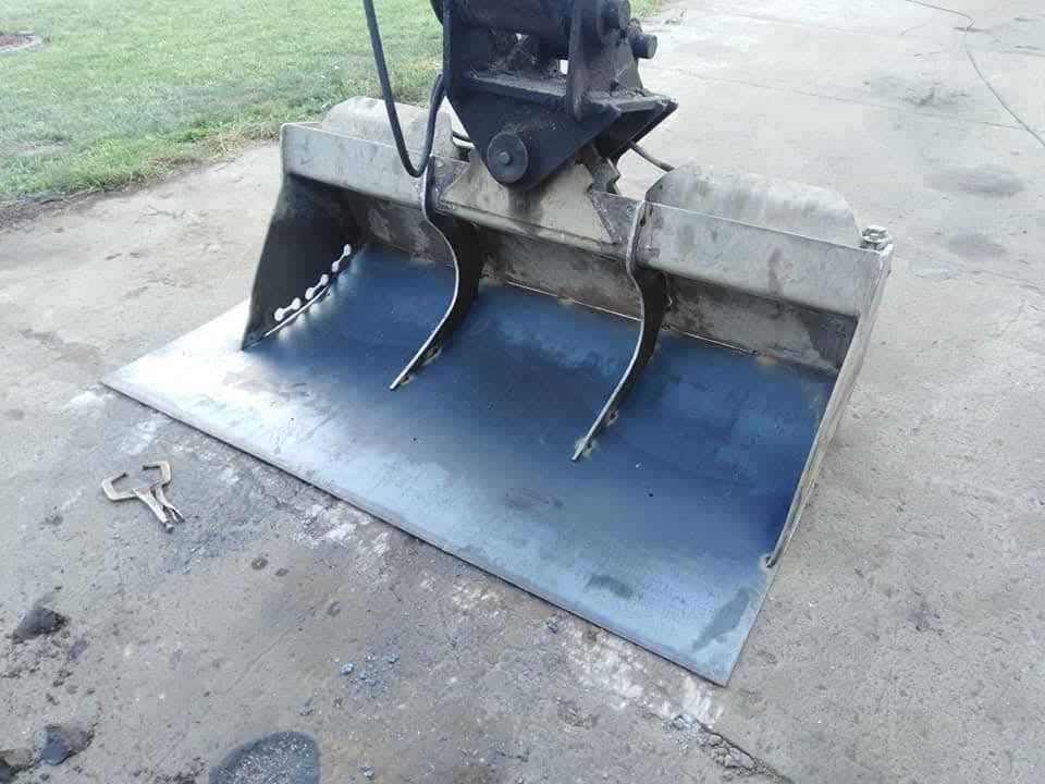 Nowo wykonana, metalowa łyżka do koparki, z widocznymi śladami spawania na gładkiej powierzchni blachy, leżąca na betonowym podłożu obok klucza hakowego.