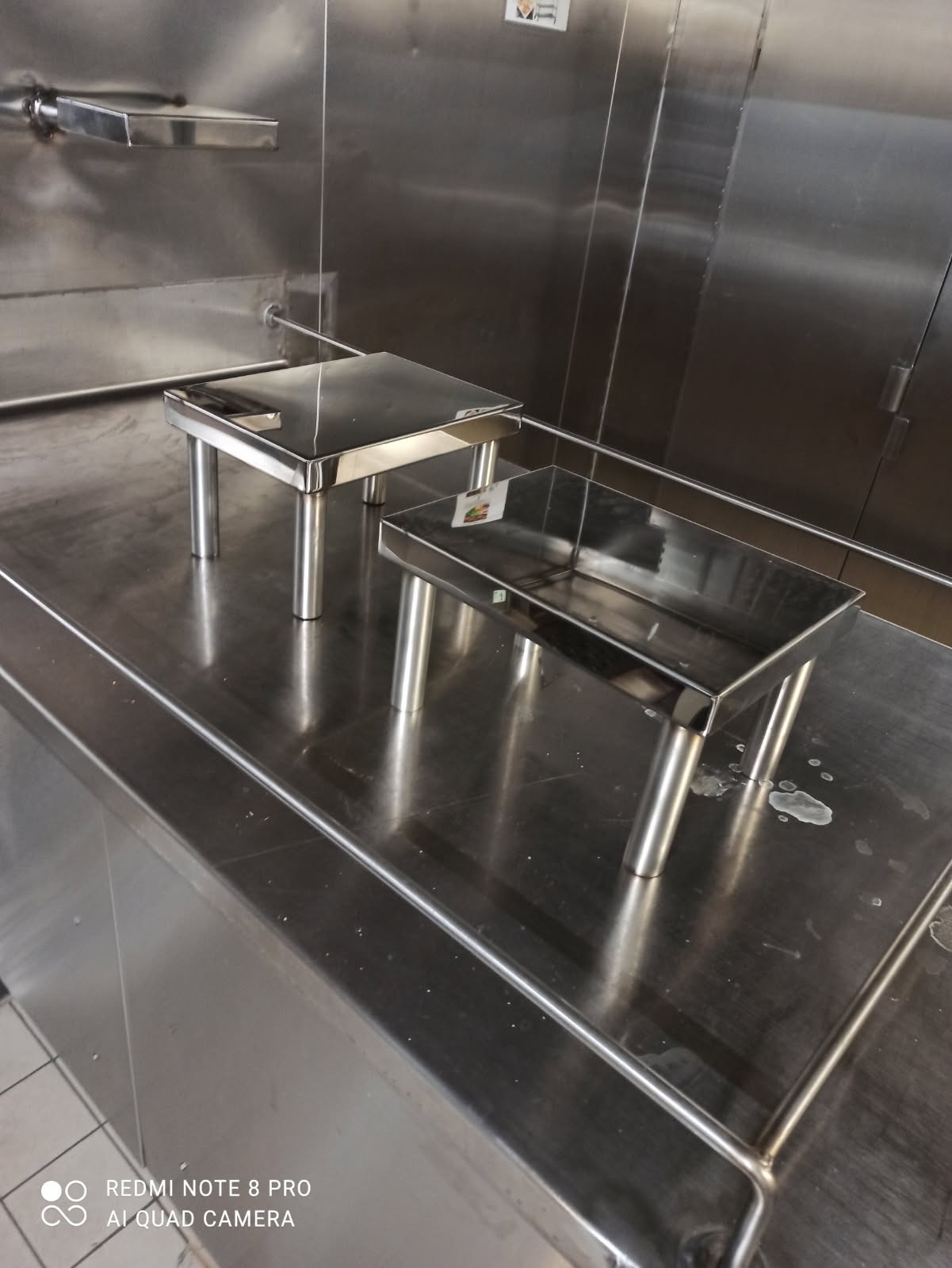 Dwie spawane, nierdzewne konstrukcje z rurek, przypominające małe stołki, stojące na nierdzewnej powierzchni.