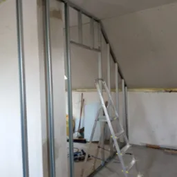 Stelaż z profili metalowych do montażu ścianki działowej z płyt gipsowo-kartonowych na poddaszu, widoczna drabina aluminiowa, narzędzia i materiały budowlane w tle.