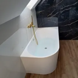 Biała wanna w łazience na poddaszu, złota bateria, ściana wyłożona czarnym marmurem z białymi żyłami, jasne panele podłogowe.