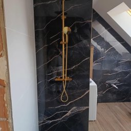 Usługi Remontowe Bez Problemu - Nowoczesna łazienka w trakcie wykańczania: złota armatura prysznicowa na tle czarnych płytek imitujących marmur z wyraźnymi złotymi żyłami, widoczne białe płytki na ścianie obok i fragment surowej...