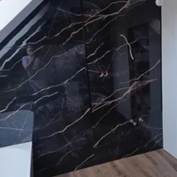 Ściana w łazience wyłożona czarnymi płytami z marmurowym wzorem, widoczne przygotowane punkty elektryczne, drewnopodobne panele podłogowe oraz fragment białej wanny.